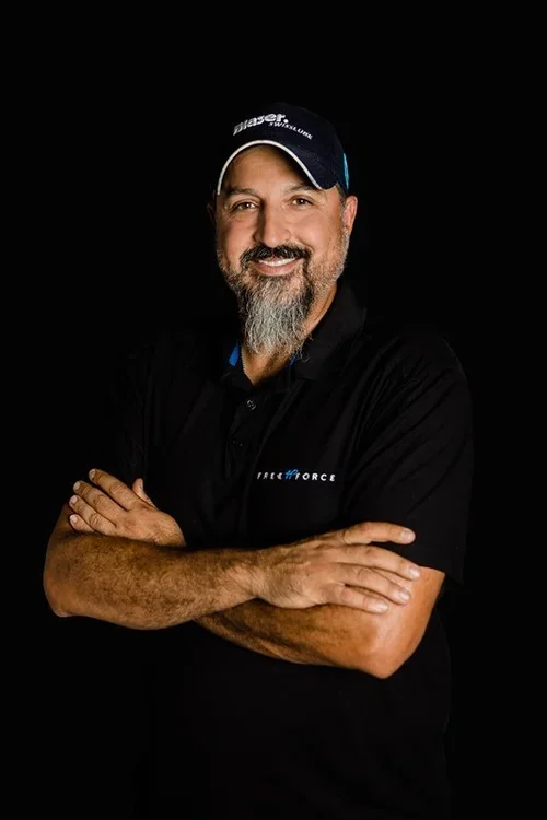 A man with a beard and mustache wearing a black polo shirt and a black cap, standing against a black background, smiling with arms crossed.