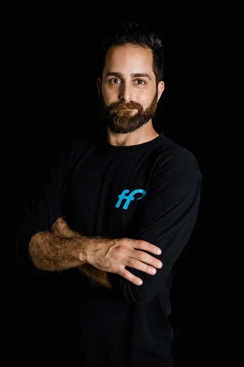 A man with dark hair and beard, wearing a black long-sleeve shirt with a blue logo, crossing his arms and standing against a black background.