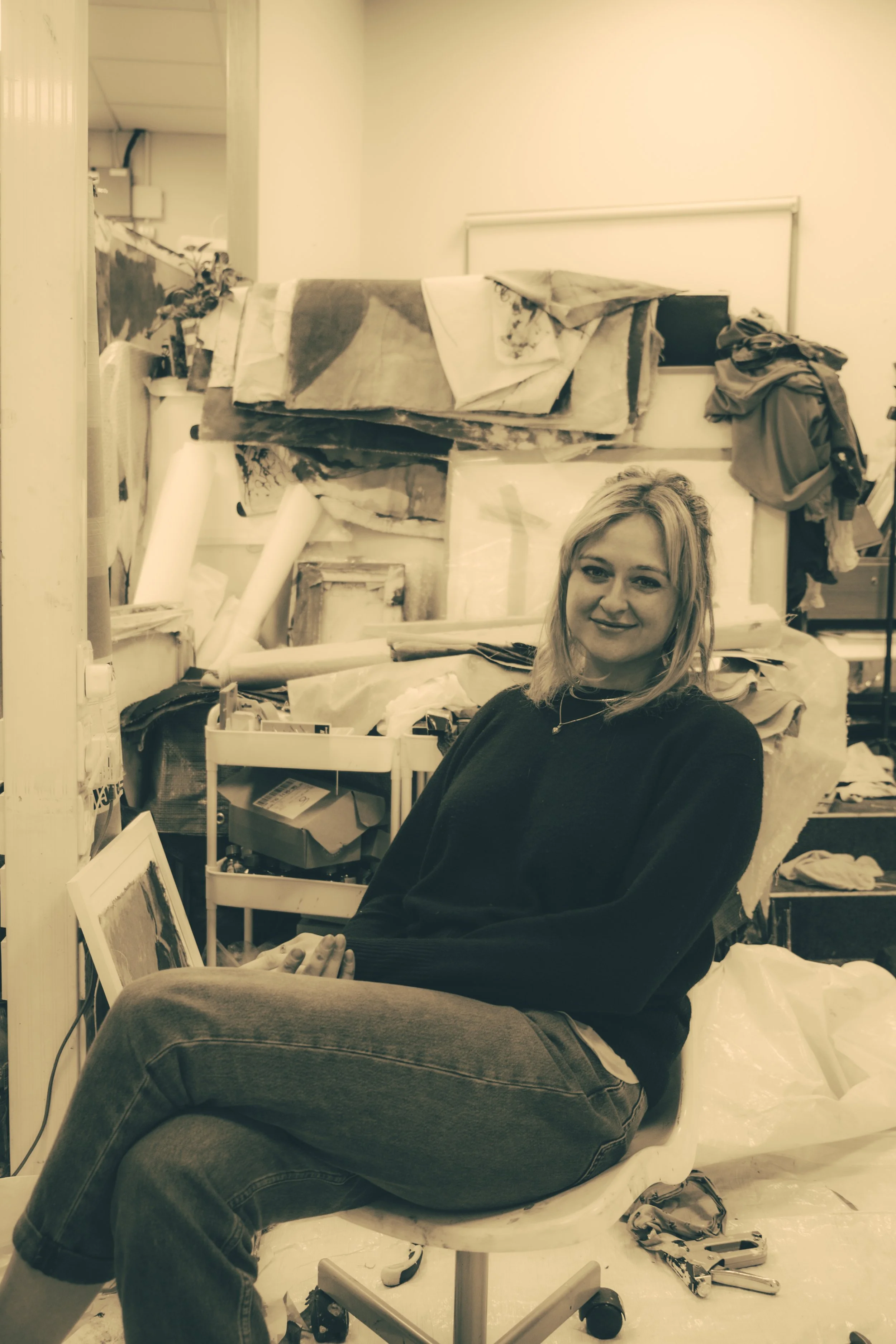 A young woman with blonde hair sitting on a white office chair surrounded by art supplies in an art studio.