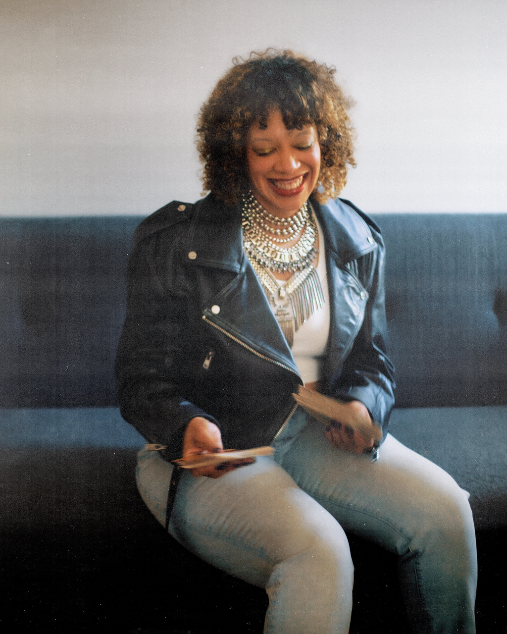 A woman with curly hair wearing a leather jacket and multiple layered pearl necklaces, smiling and sitting on a dark couch, holding playing cards.