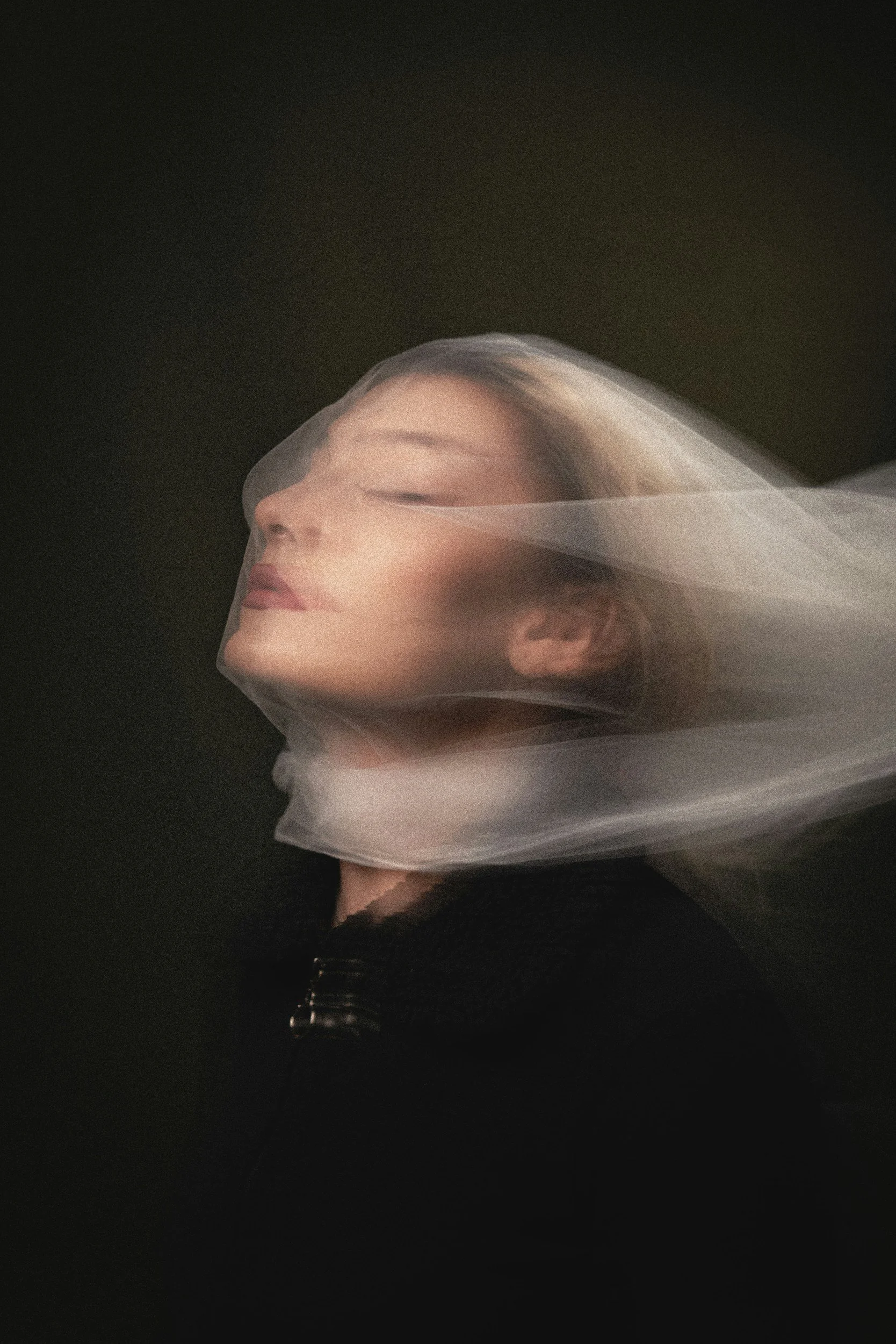 A woman with blonde hair and makeup, her eyes closed, is wearing a sheer veil that covers her head and flows downward, set against a dark background.