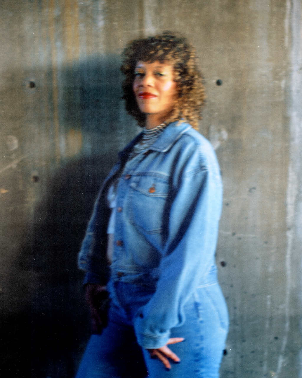 A woman with curly hair wearing a denim jacket and striped t-shirt, standing against a concrete wall.