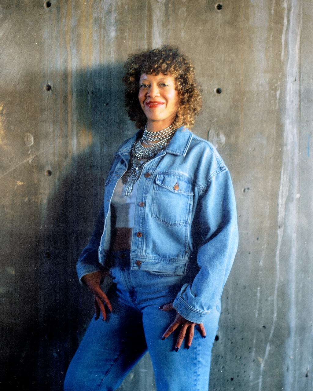 A woman with curly hair wearing a denim jacket and jeans, accessorized with multiple necklaces, standing against a concrete wall.