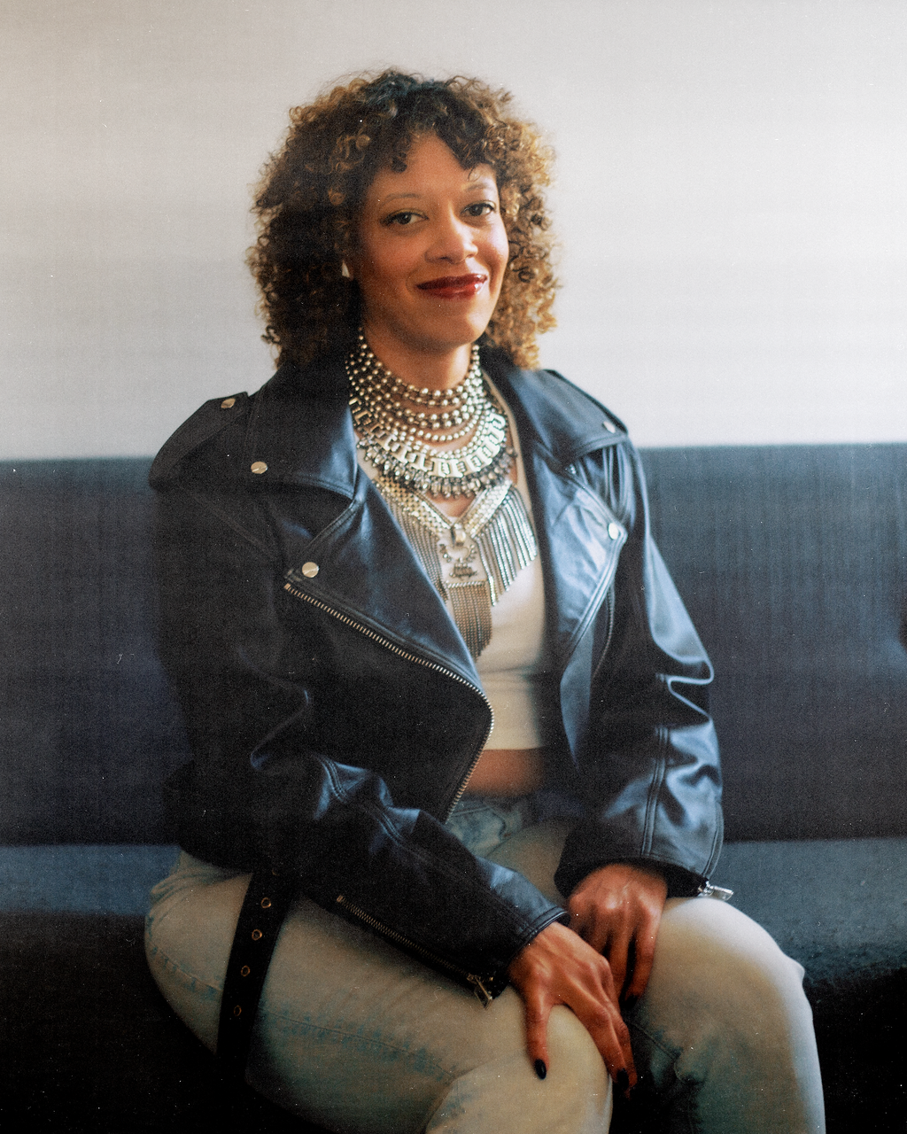 A woman with curly hair sitting on a dark-colored couch, wearing a black leather jacket, white crop top, and multiple layered pearl and metallic necklaces, smiling at the camera.