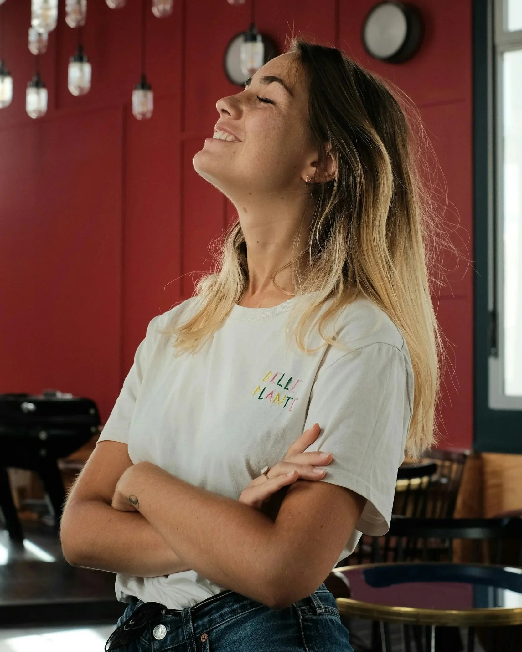 Woman with eyes closed and head tilted upward, standing with arms crossed in front of a red wall with hanging lights.