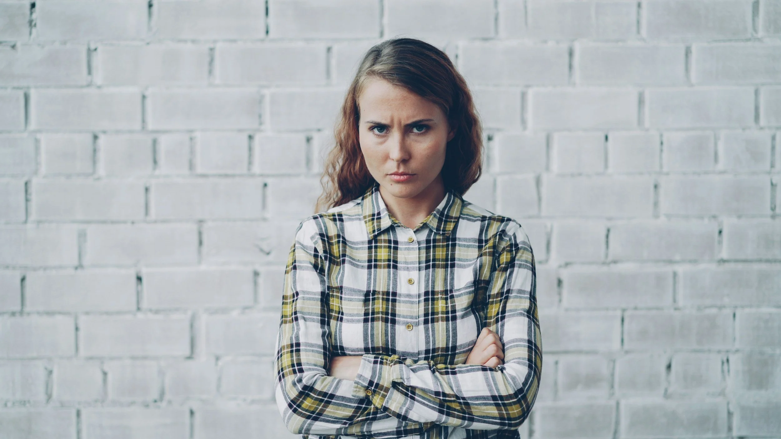 A woman with a serious expression stands against a white brick wall with her arms crossed, conveying frustration or defensiveness.