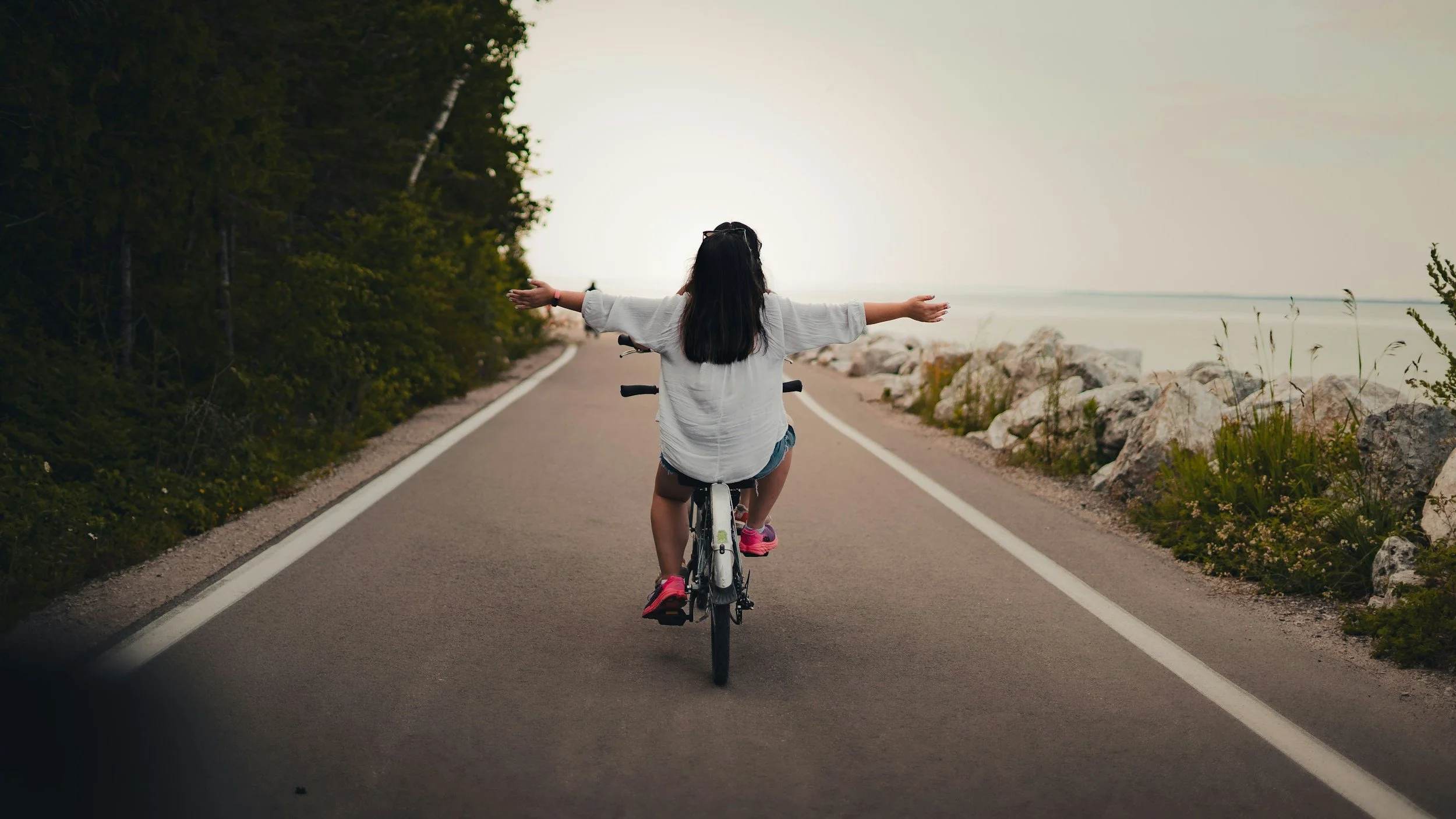A person rides a bicycle down a quiet road with arms outstretched, expressing freedom and balance in an open outdoor setting.