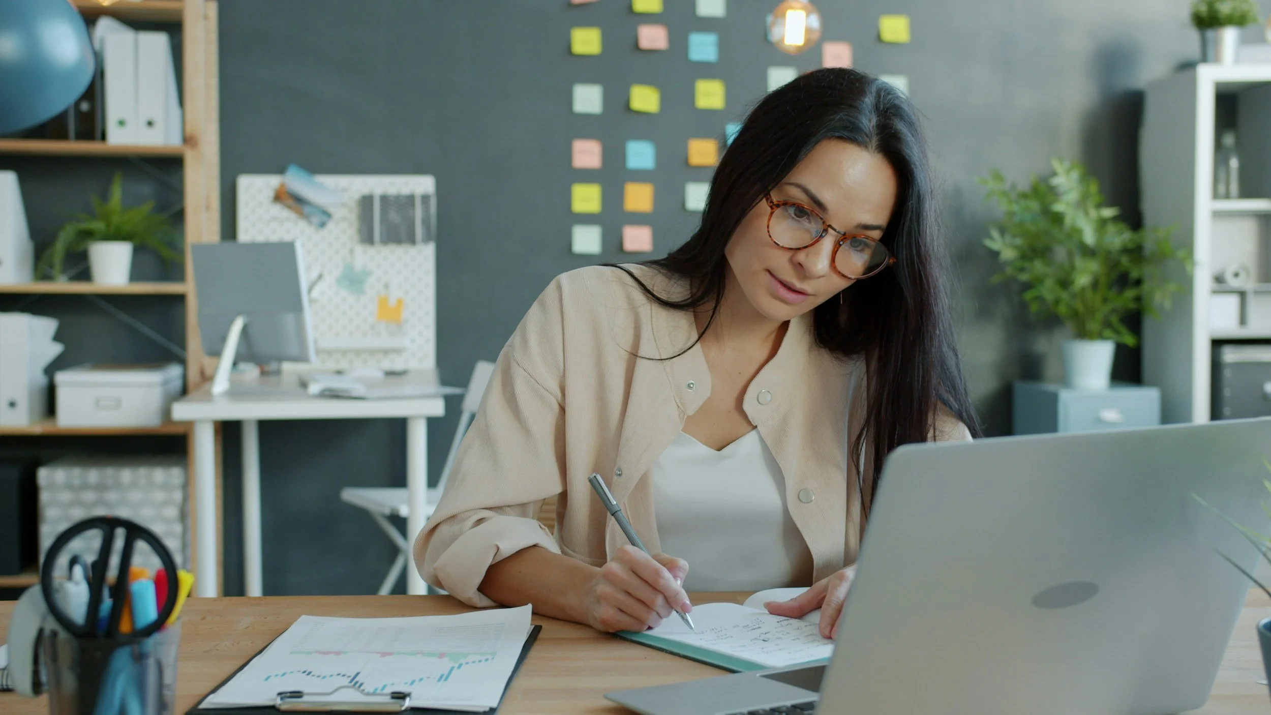 Woman working at a desk, writing in a notebook while using a laptop in a calm, organized workspace