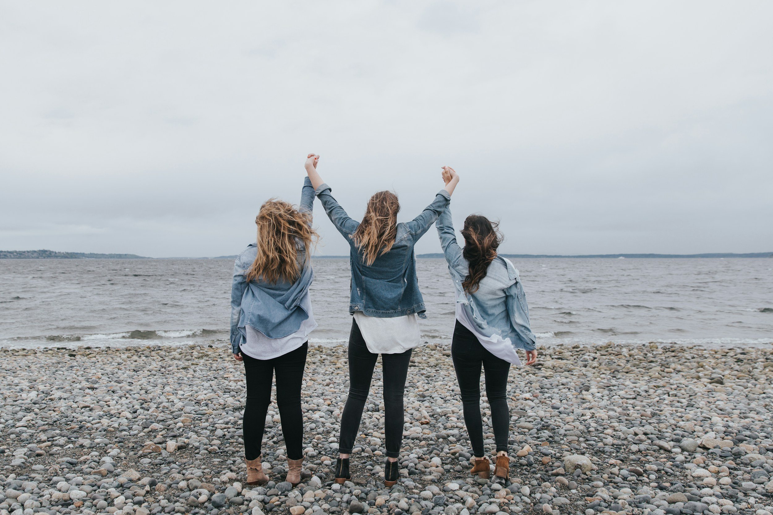 Three women seen from behind, holding hands raised in the air on a rocky beach.