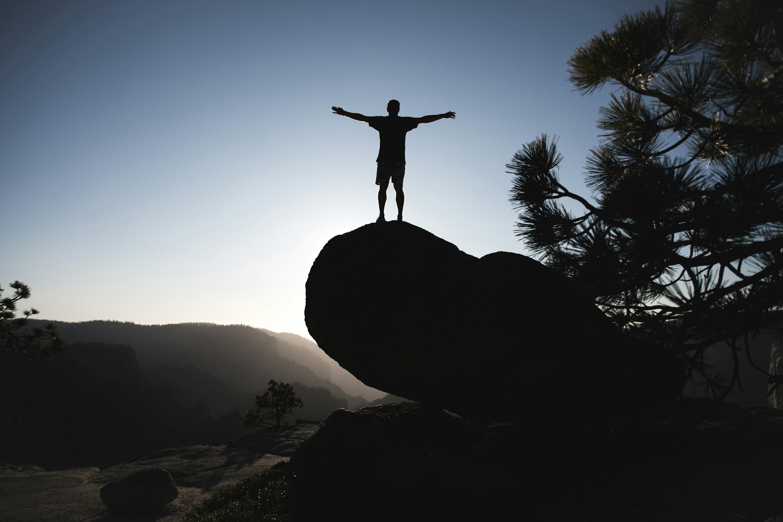 Person standing on a large rock with arms outstretched overlooking a mountain landscape.