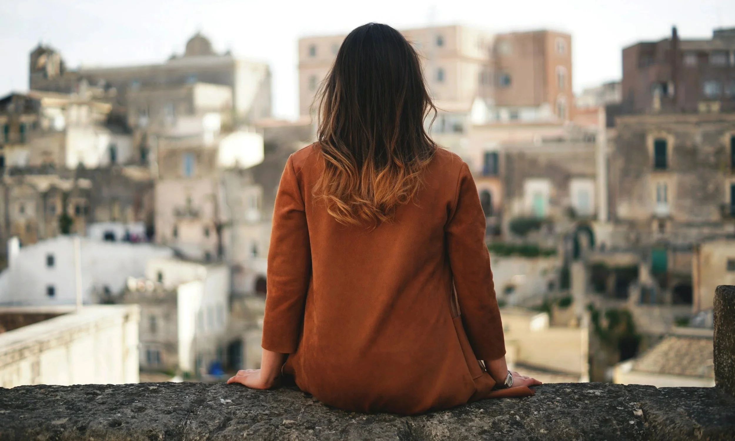 Person sitting on a stone wall overlooking a city, reflecting quietly and looking outward