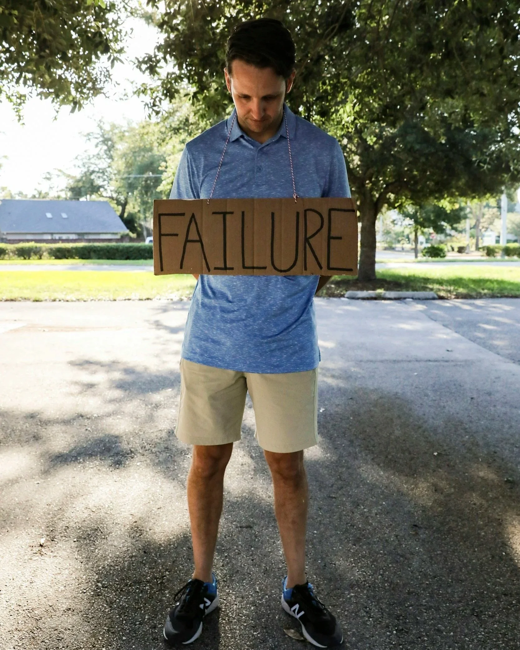 Man standing outdoors with head down wearing a cardboard sign that says “Failure,” expressing feelings of shame or self-doubt.