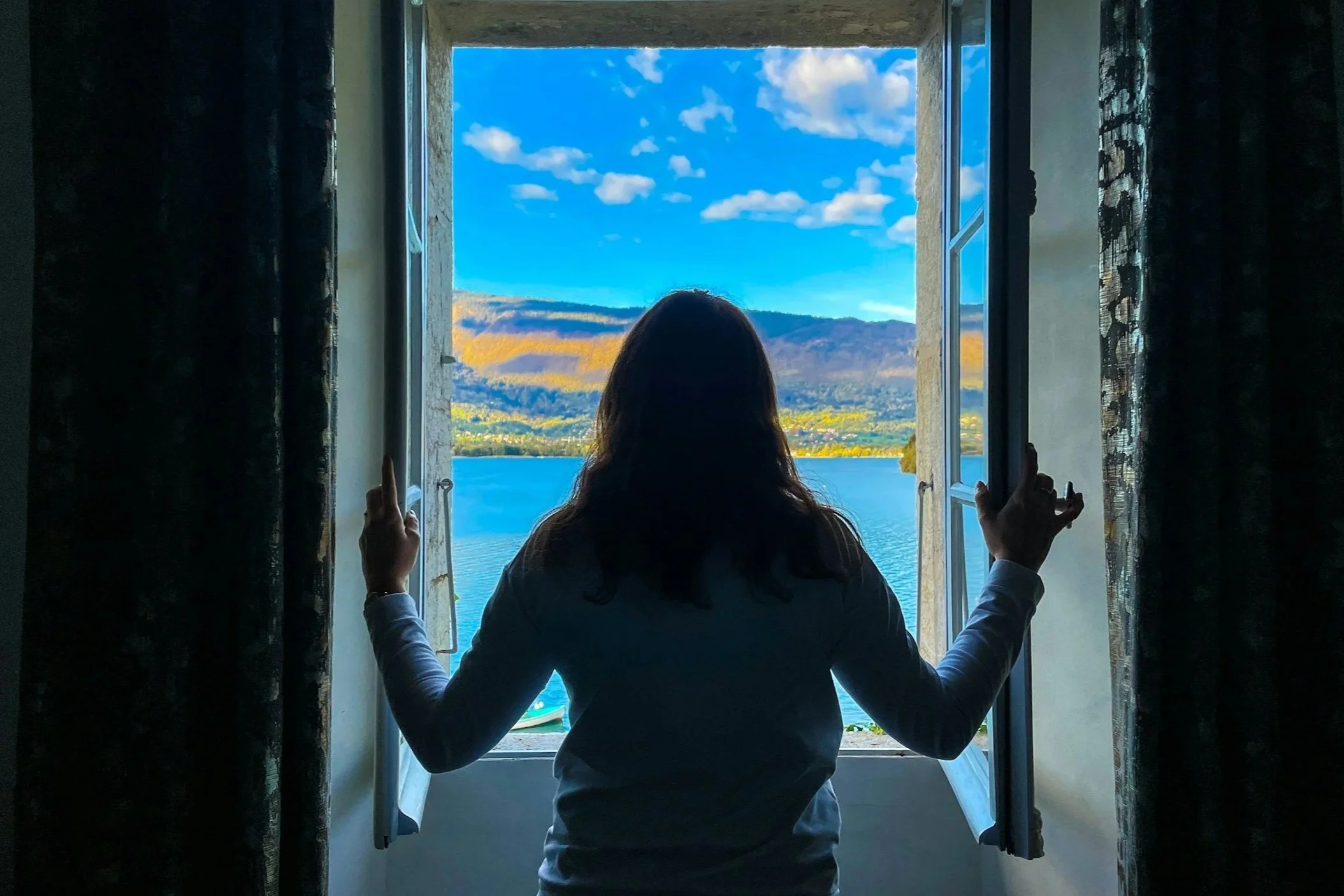 Person standing with their back to the camera, holding open two windows overlooking a blue lake and mountains.