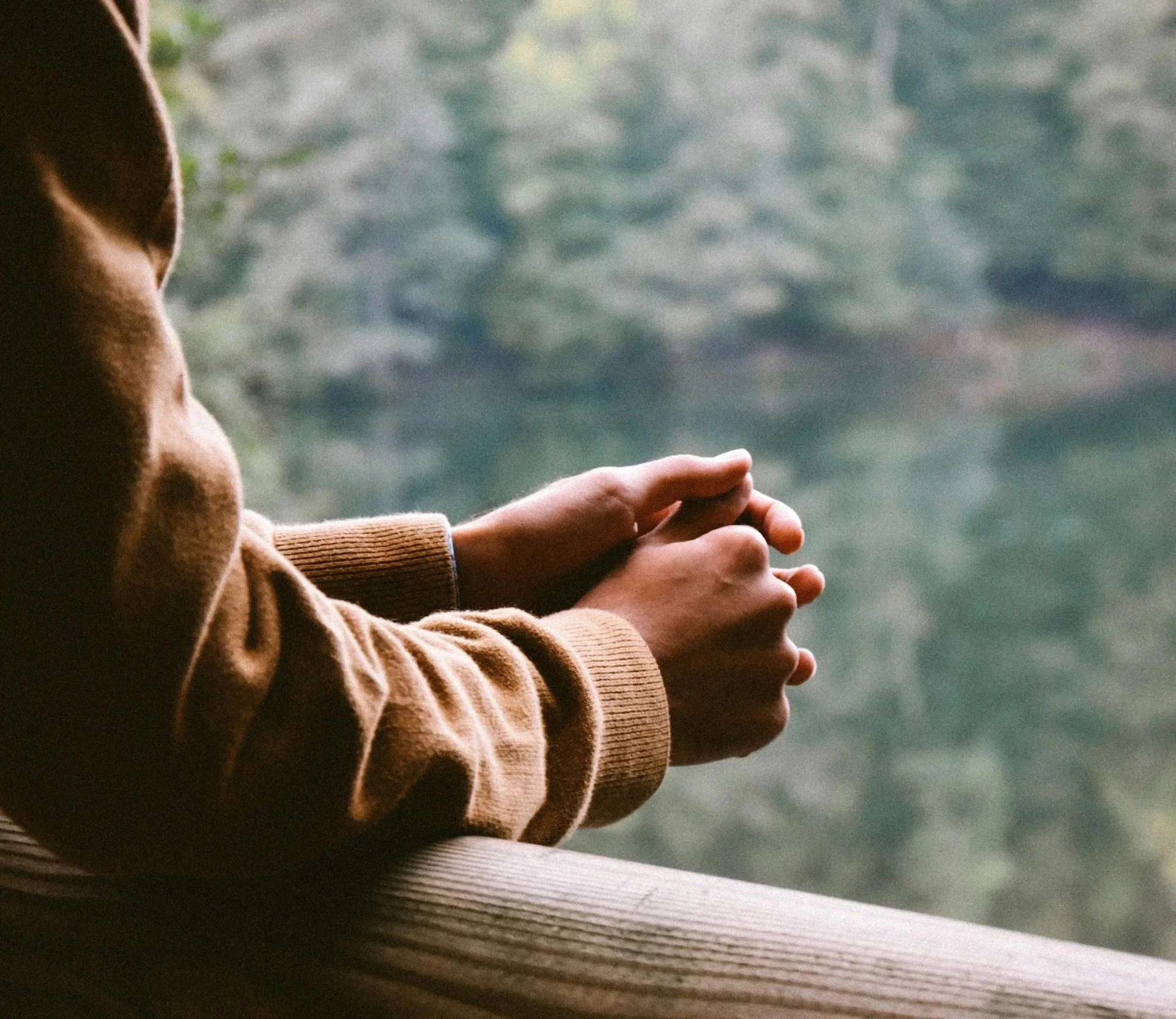 person's arms resting on a wooden railing with hands clasped, overlooking water and trees