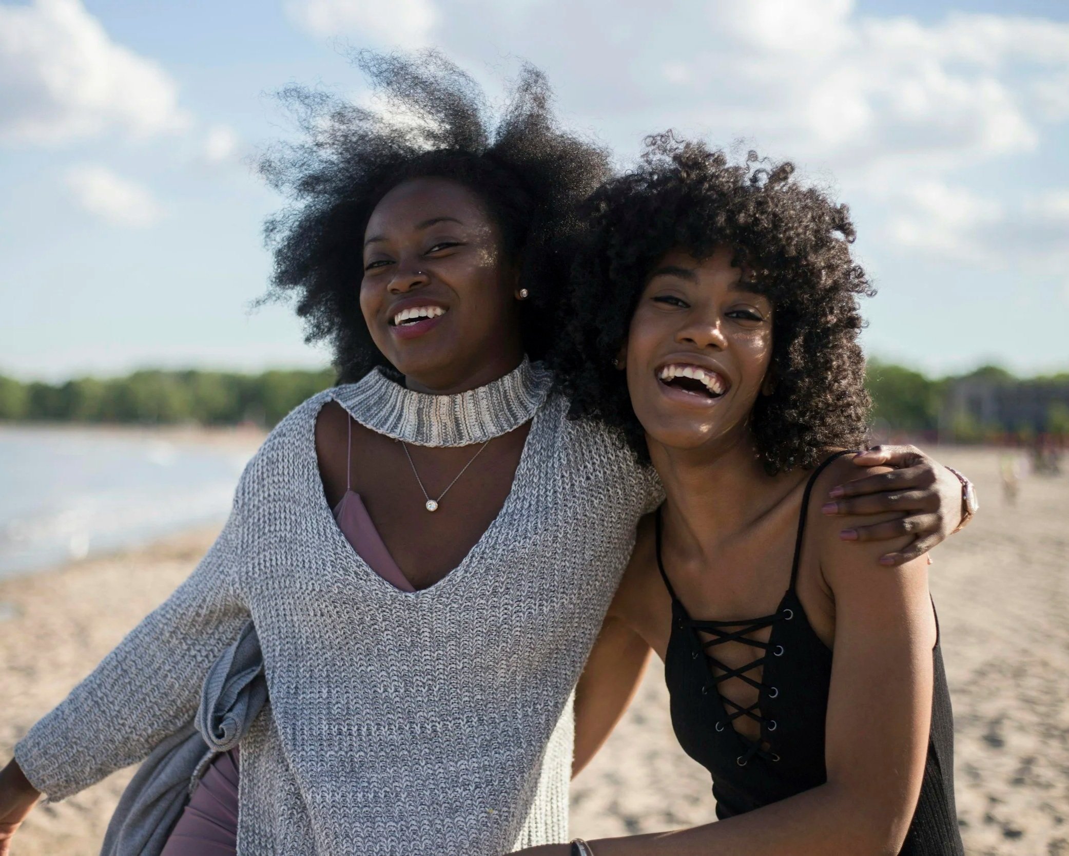 Two women smile and laugh together on a sunny beach, standing close with arms around each other, conveying connection and joy.
