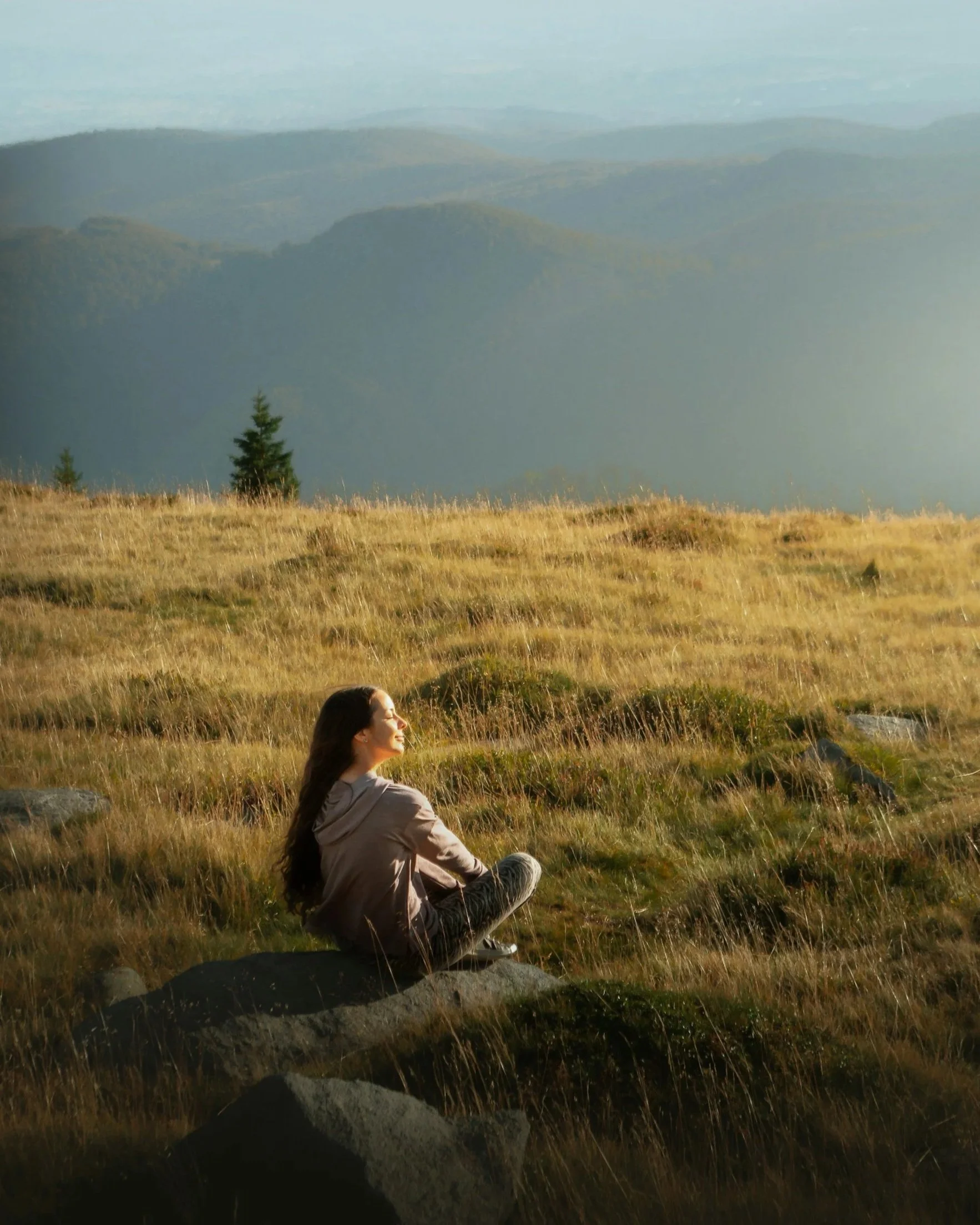 A woman sits on a rock in an open grassy landscape, looking out over distant mountains, conveying calm, reflection, and spaciousness.