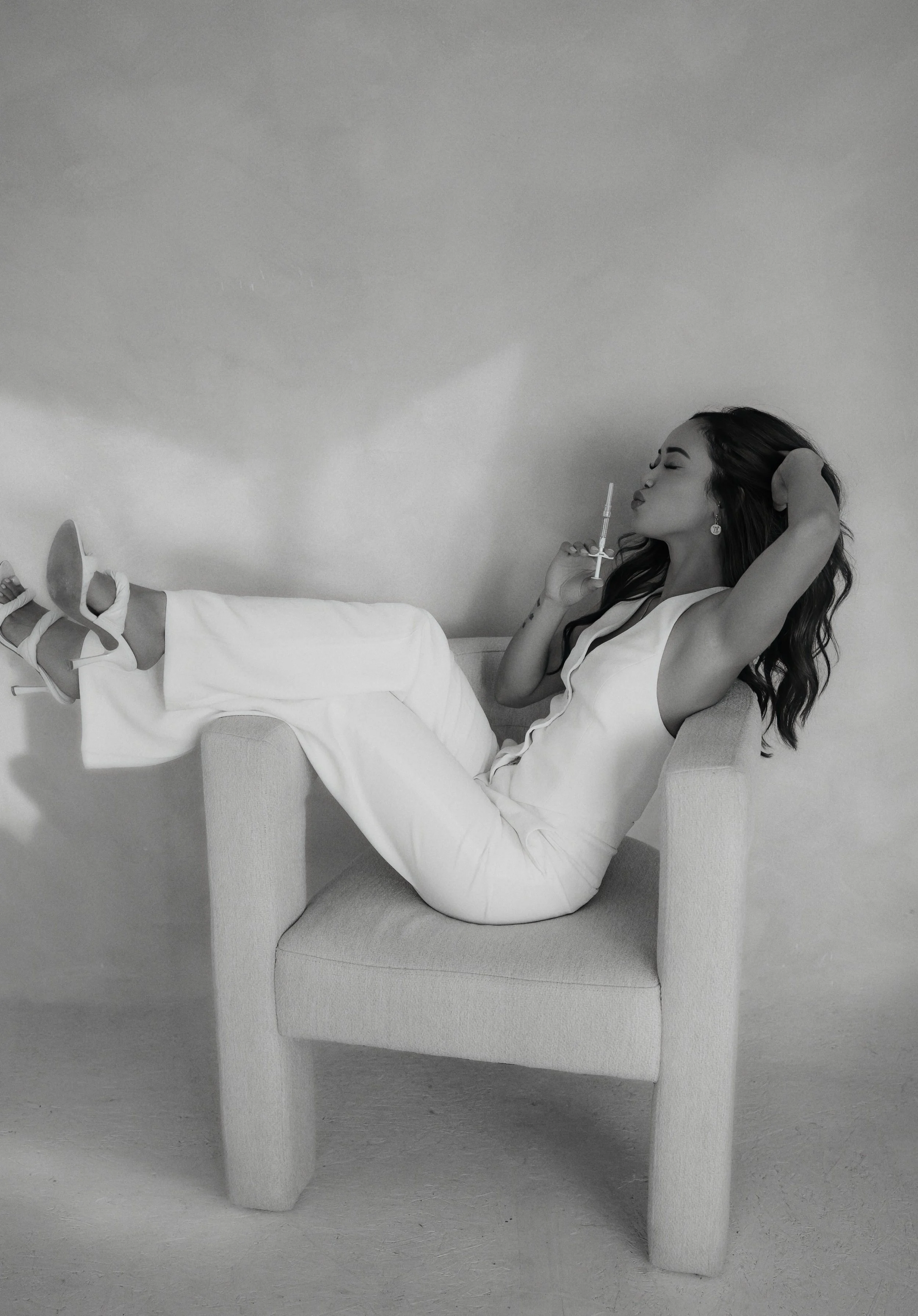 A woman in white clothing reclining on a chair, holding a syringe close to her lips, with high heels on her feet and one arm behind her head, in black and white.