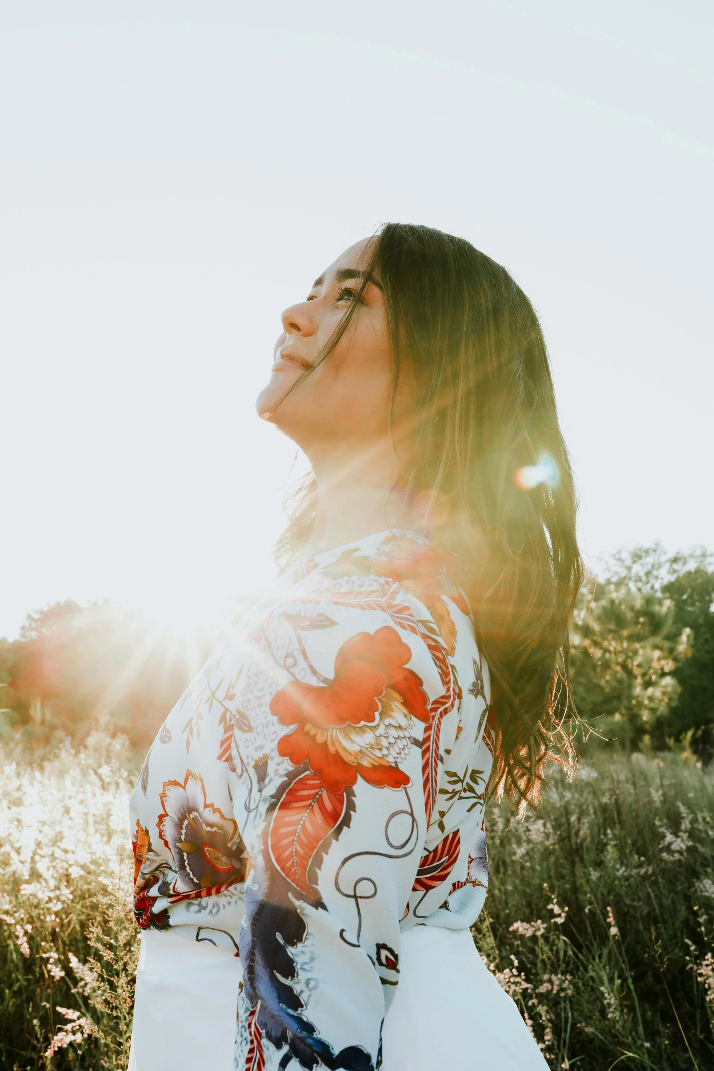A woman with long dark hair looking upwards, standing in a field with sunlight behind her on a clear day.