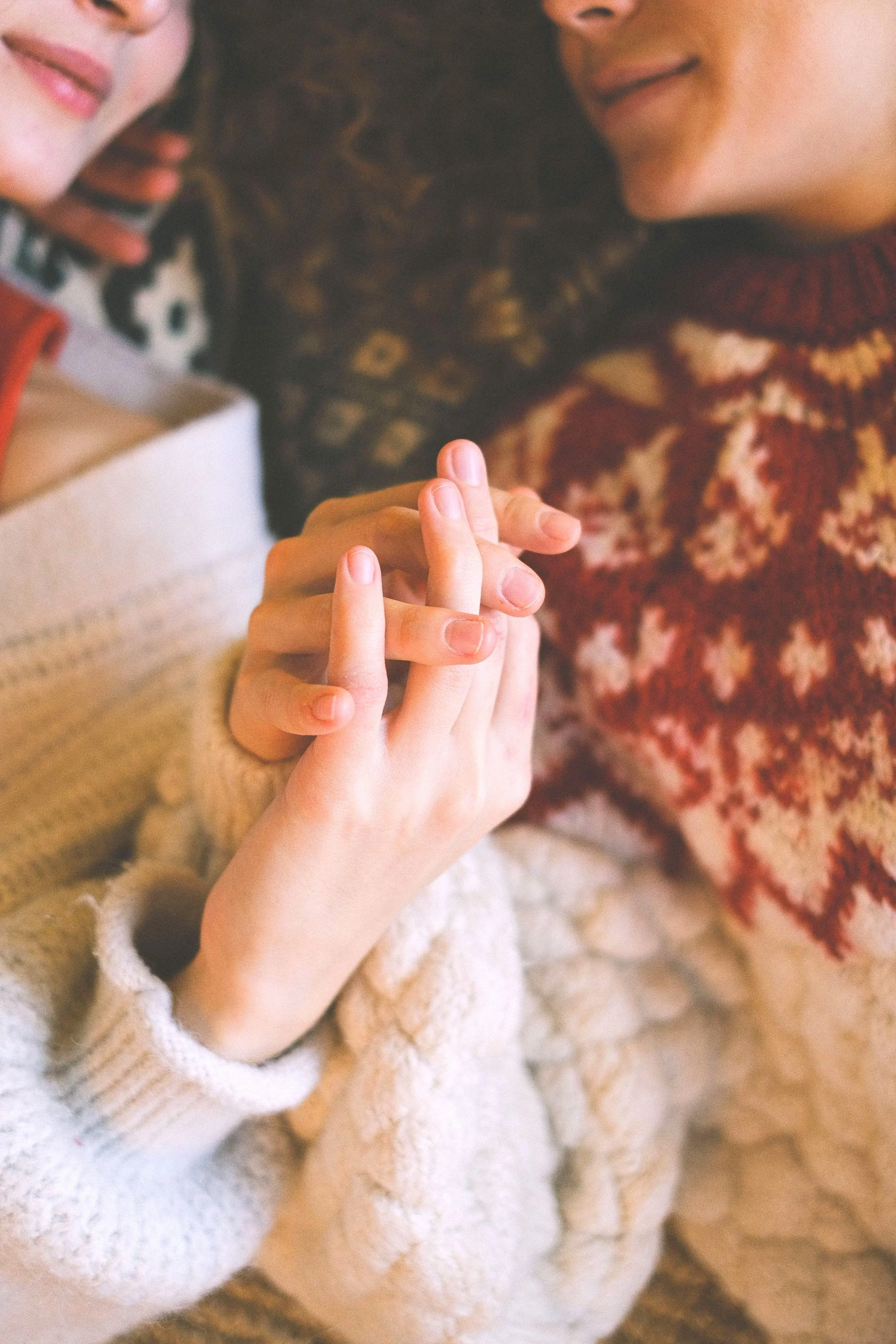 Two children holding hands, one wearing a beige coat and the other wearing a patterned sweater with red, orange, and beige colors. Focus on their hands and part of their faces.