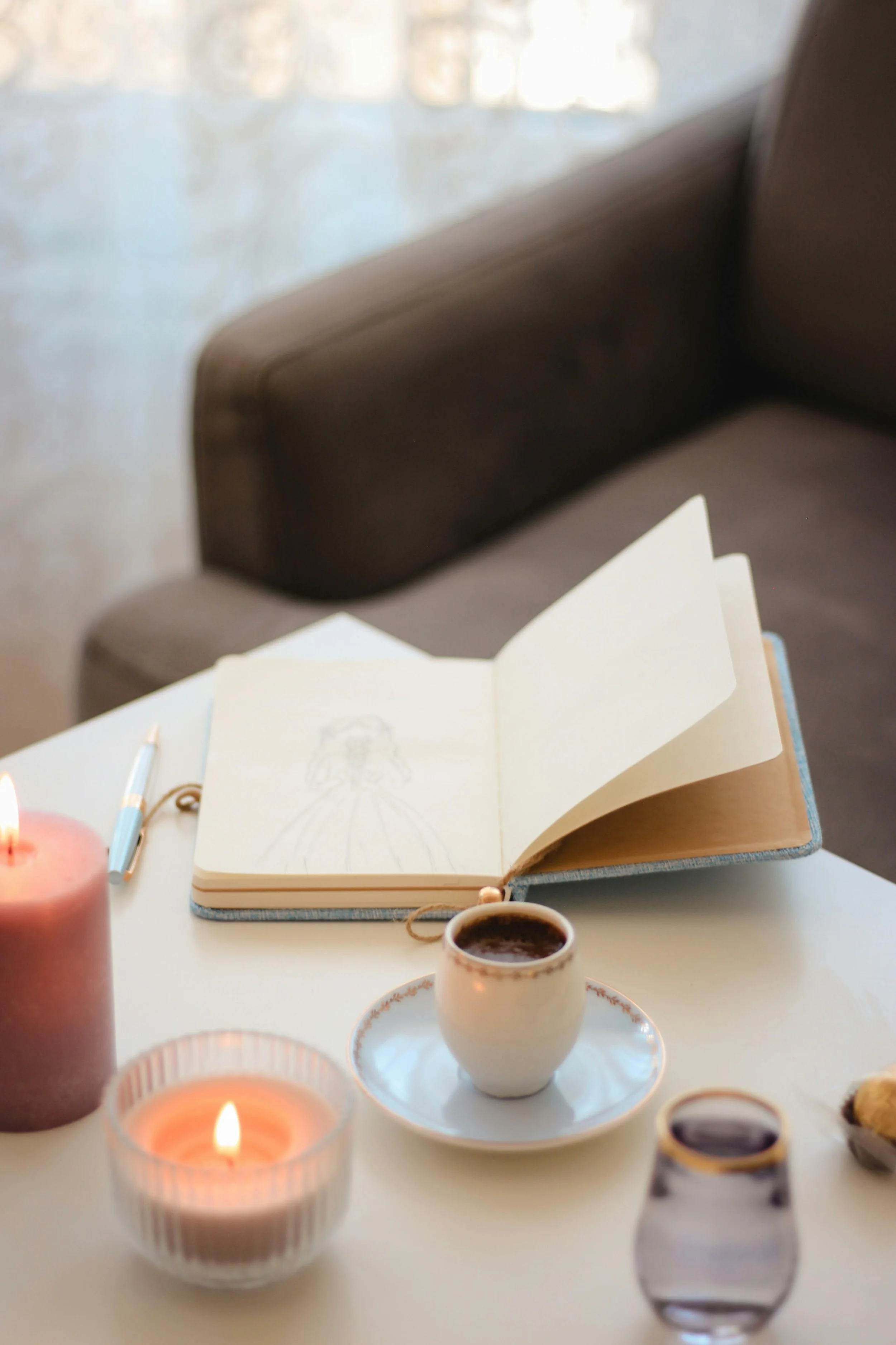 Open sketchbook with a pencil on a table surrounded by candles, cups of coffee, and decorative items.