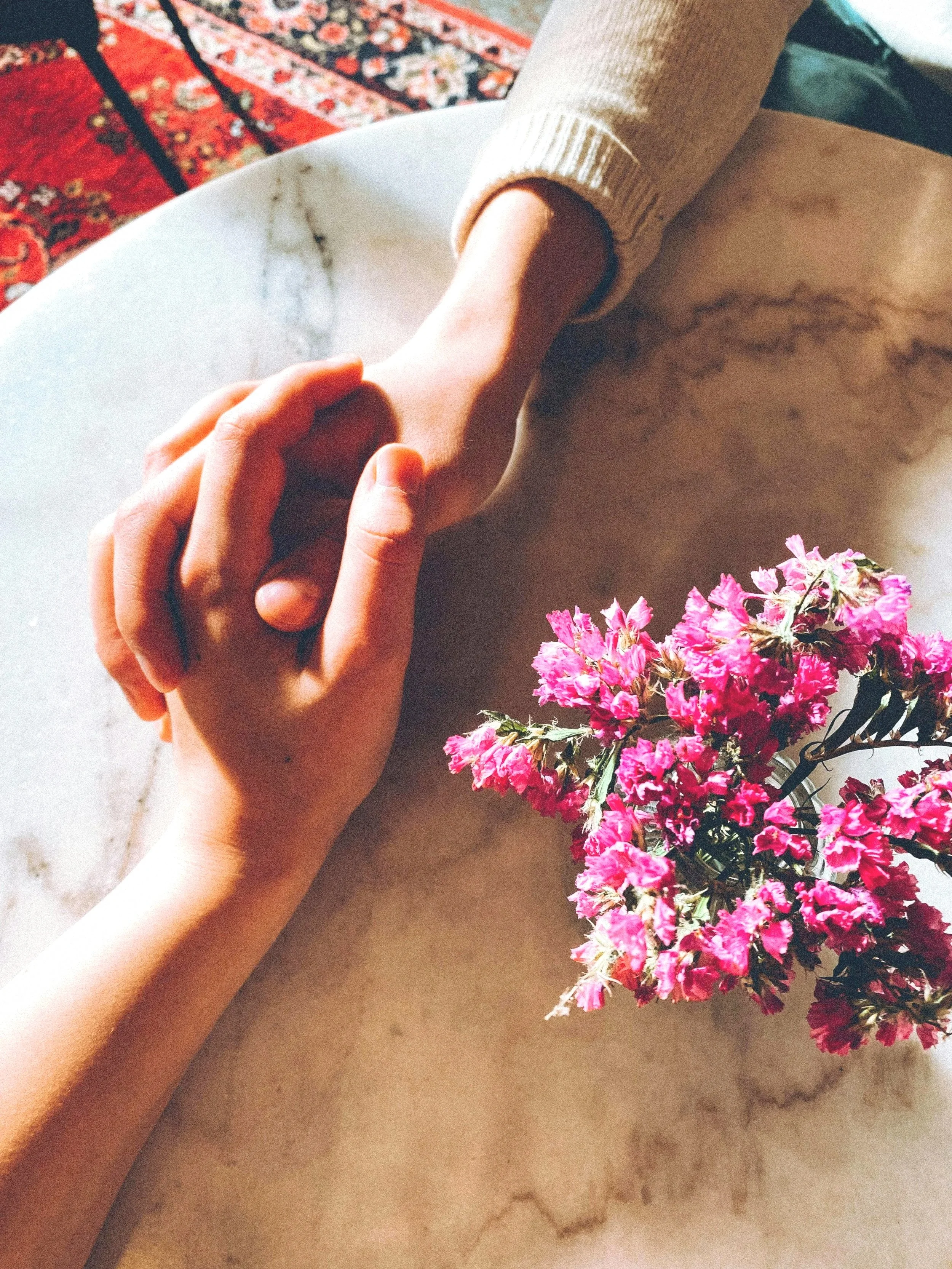 A person holding hands with another arm, with pink flowers on a marble surface, a white table, and a patterned red and black carpet in the background.