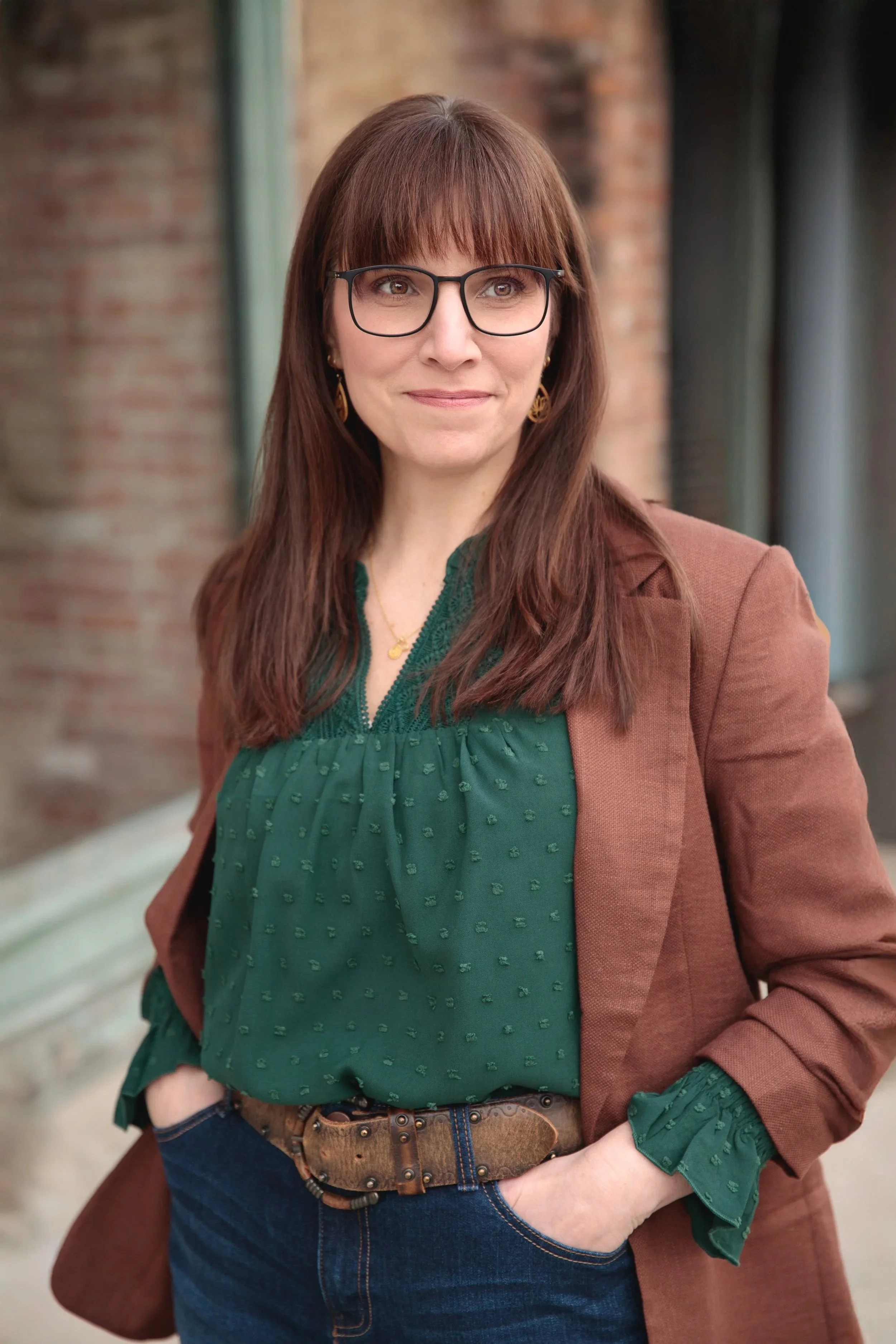 A woman with brown hair, glasses, and earrings wearing a green blouse and a brown blazer, standing outdoors with a brick wall and windows in the background.