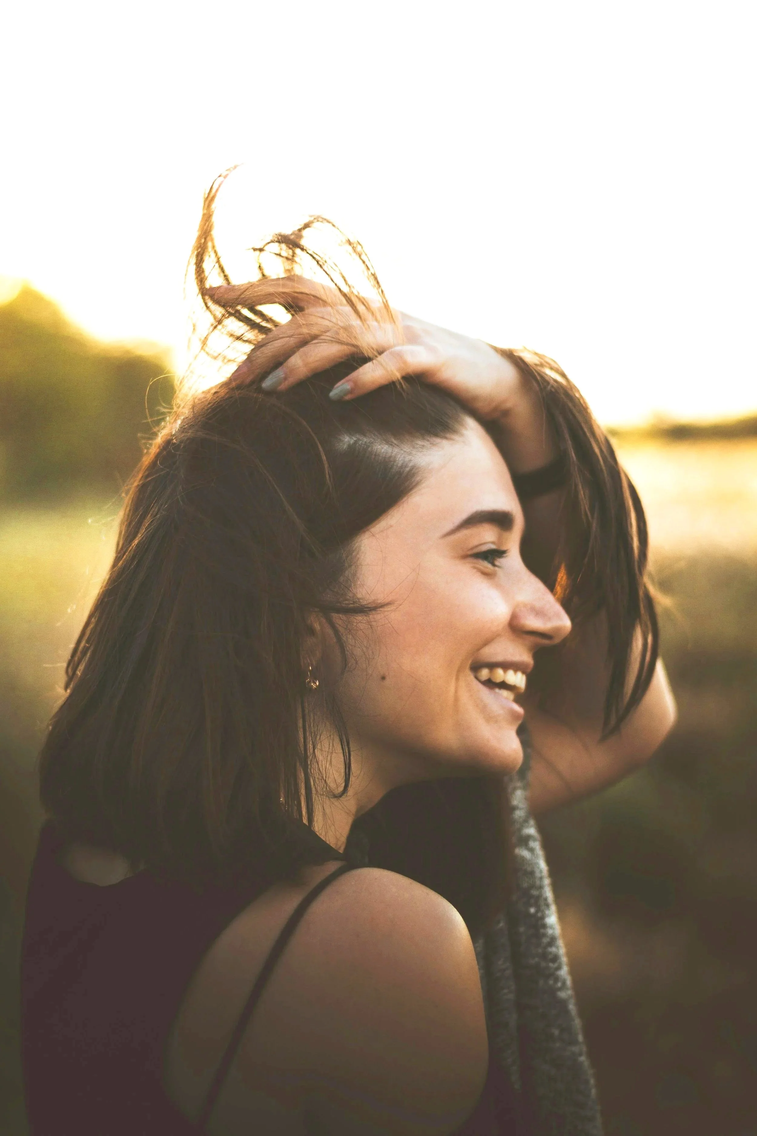 Young woman with long dark hair, smiling outdoors during sunset, wearing a sleeveless top, with her hand in her hair.