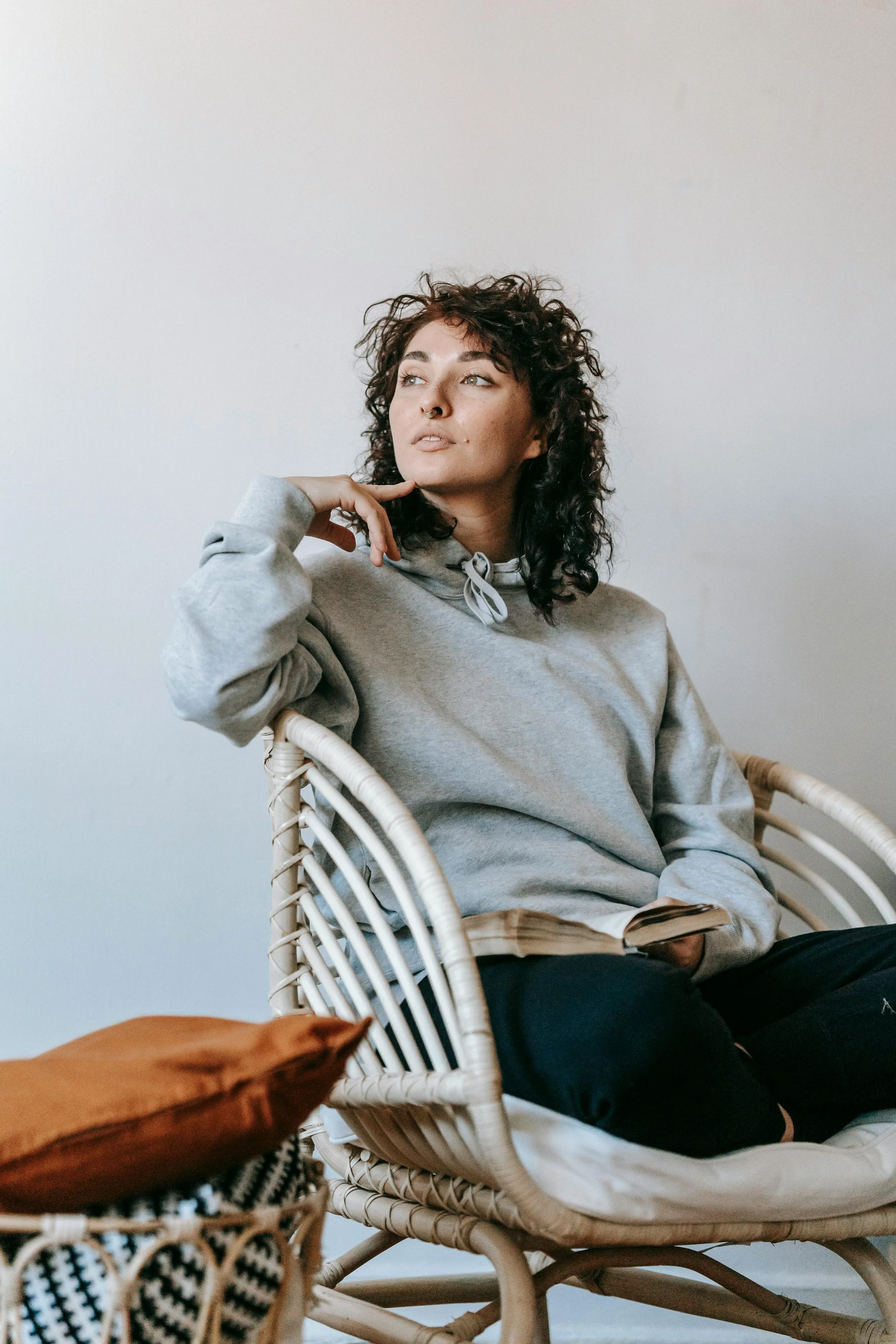 Young woman with curly dark hair wearing a gray hoodie, sitting in a rattan chair, looking thoughtfully to her left. She has a septum piercing and is in a room with plain white walls.