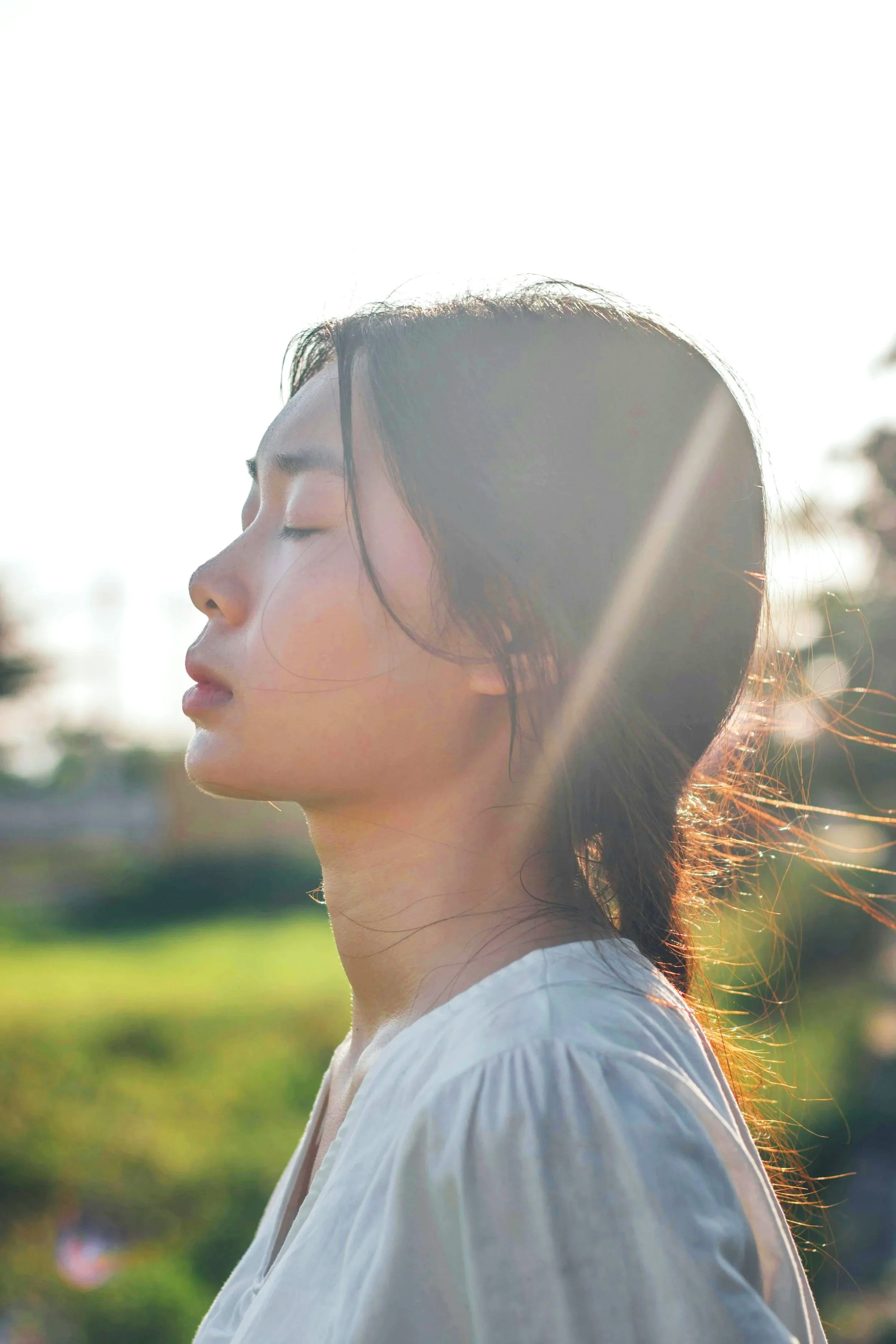 Profile of a young woman with closed eyes, dark hair, and light skin, outdoors in sunlight, wearing a white shirt.