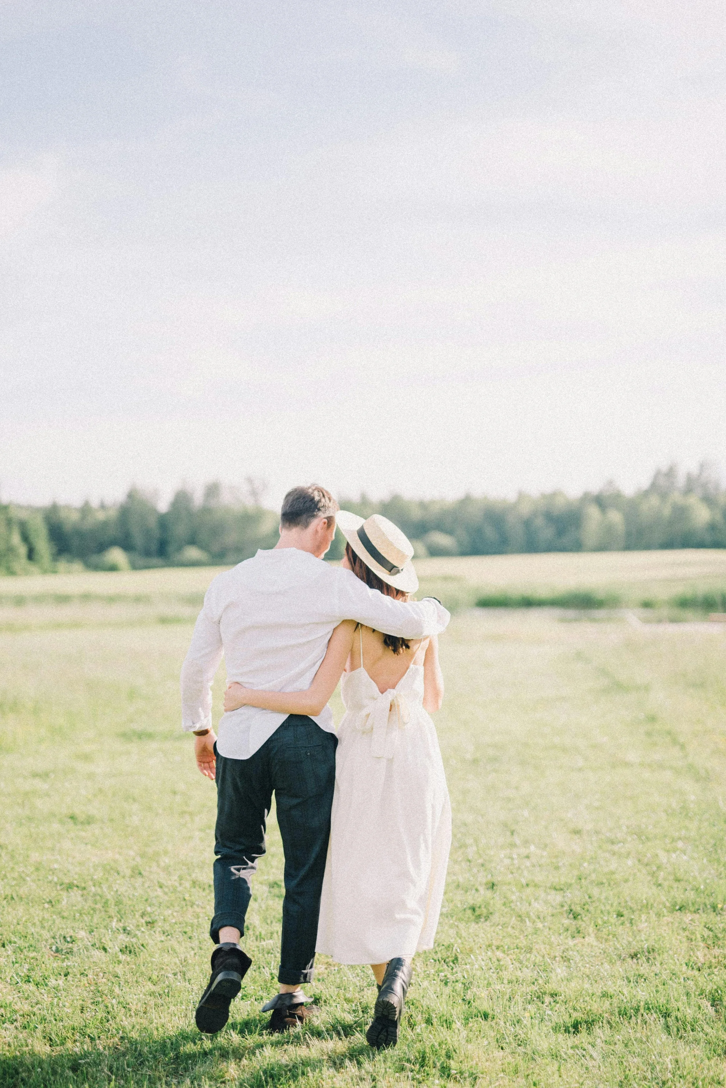 A couple walking in a grassy field on a sunny day, with the man wearing a white shirt and dark pants and the woman wearing a white dress and a straw hat.