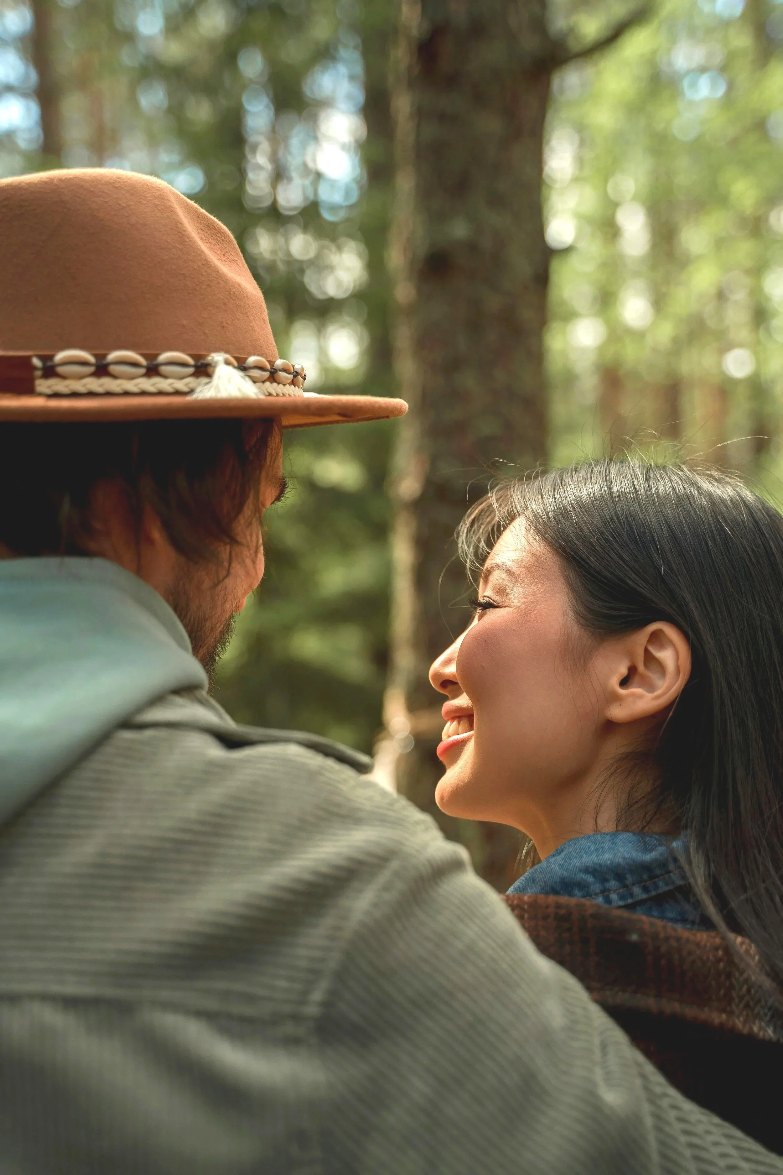 A man and a woman smiling and looking into each other's eyes in a forest setting, with the man's face mostly obscured by a hat and the woman's face visible in profile.
