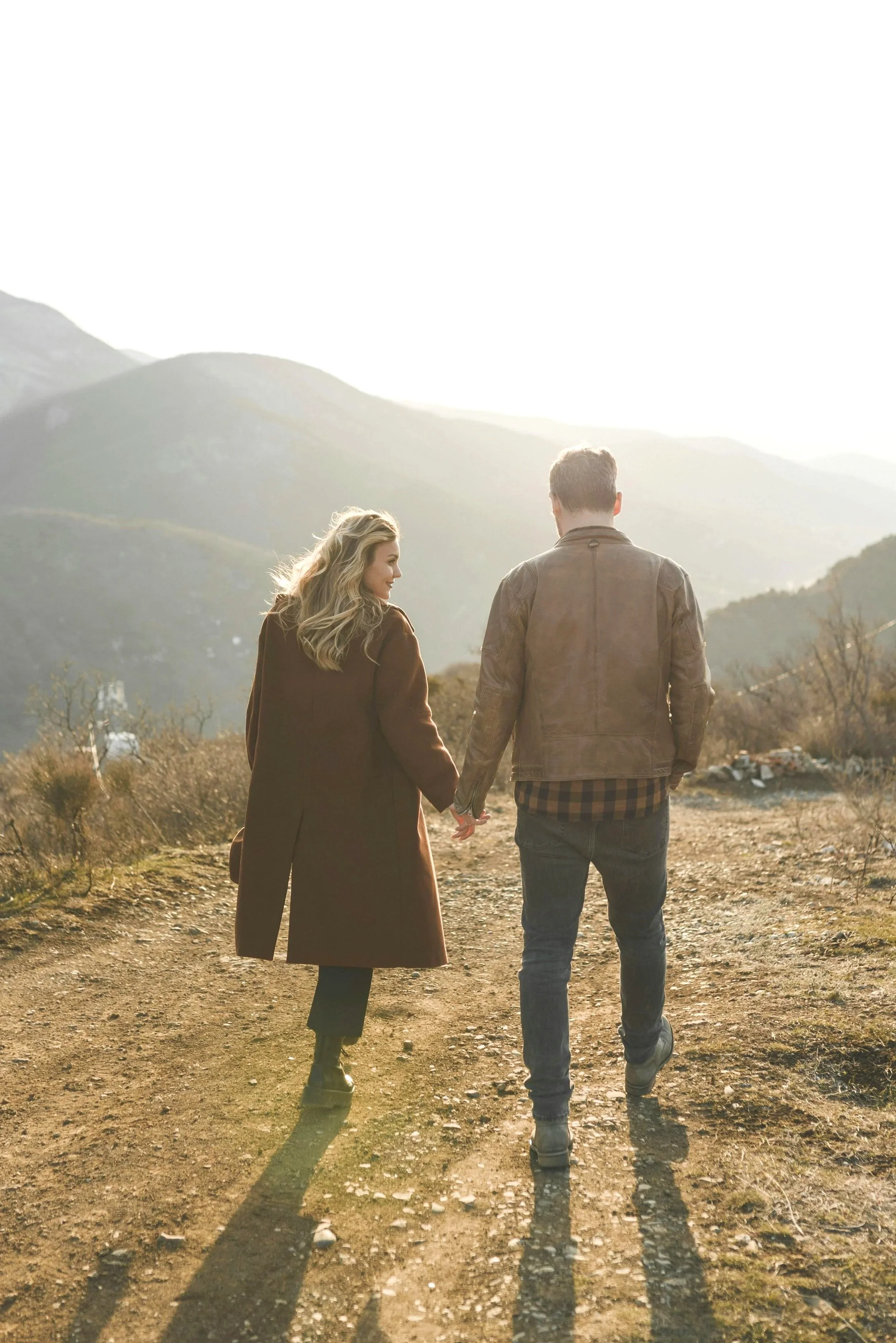 A young couple walking hand-in-hand outdoors on a dirt path with mountains in the background during sunset, dressed casually and holding hands.