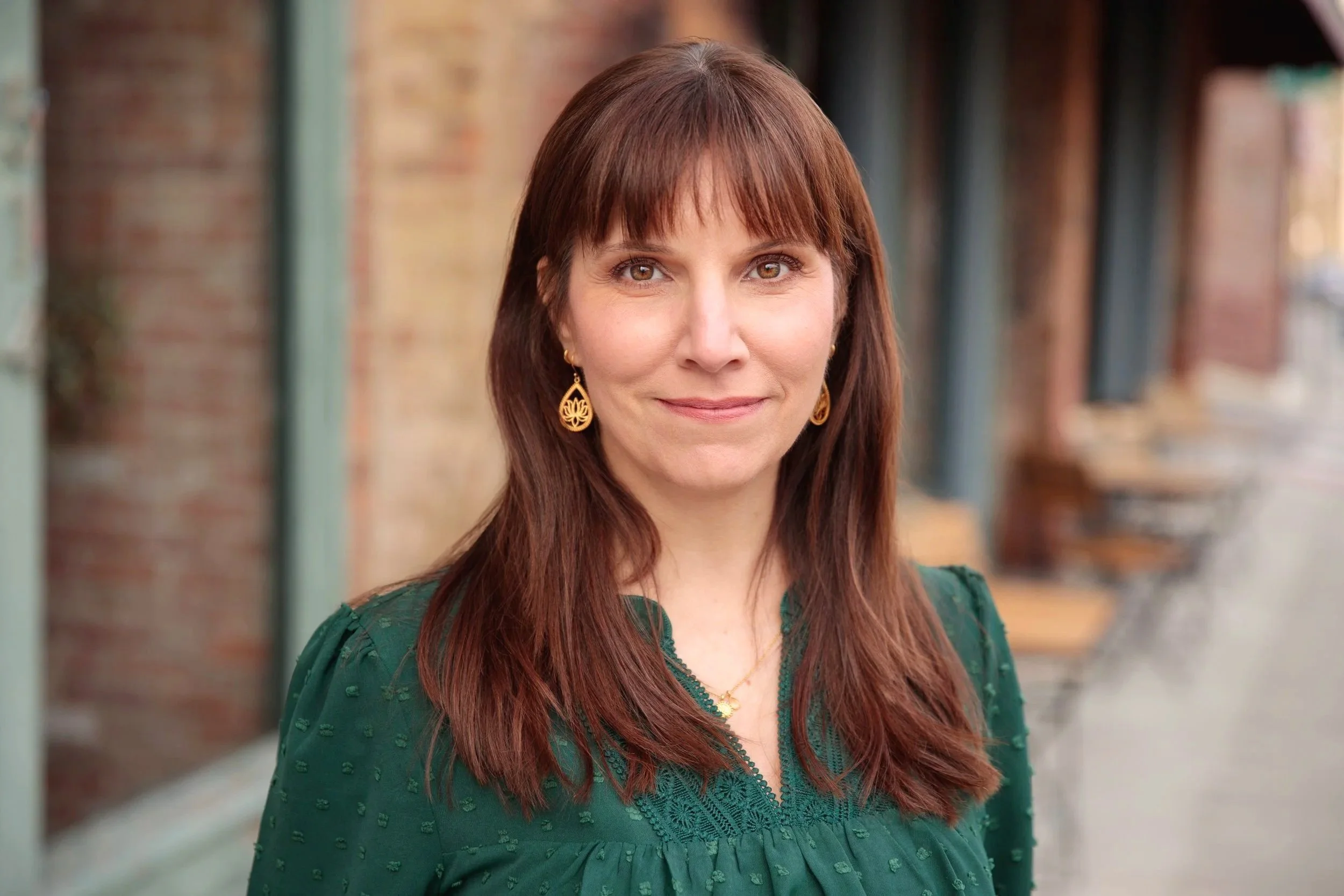 A woman with long brown hair and light brown eyes is smiling, wearing gold earrings and a green embroidered top, standing outdoors with blurred brick walls and pillars in the background.