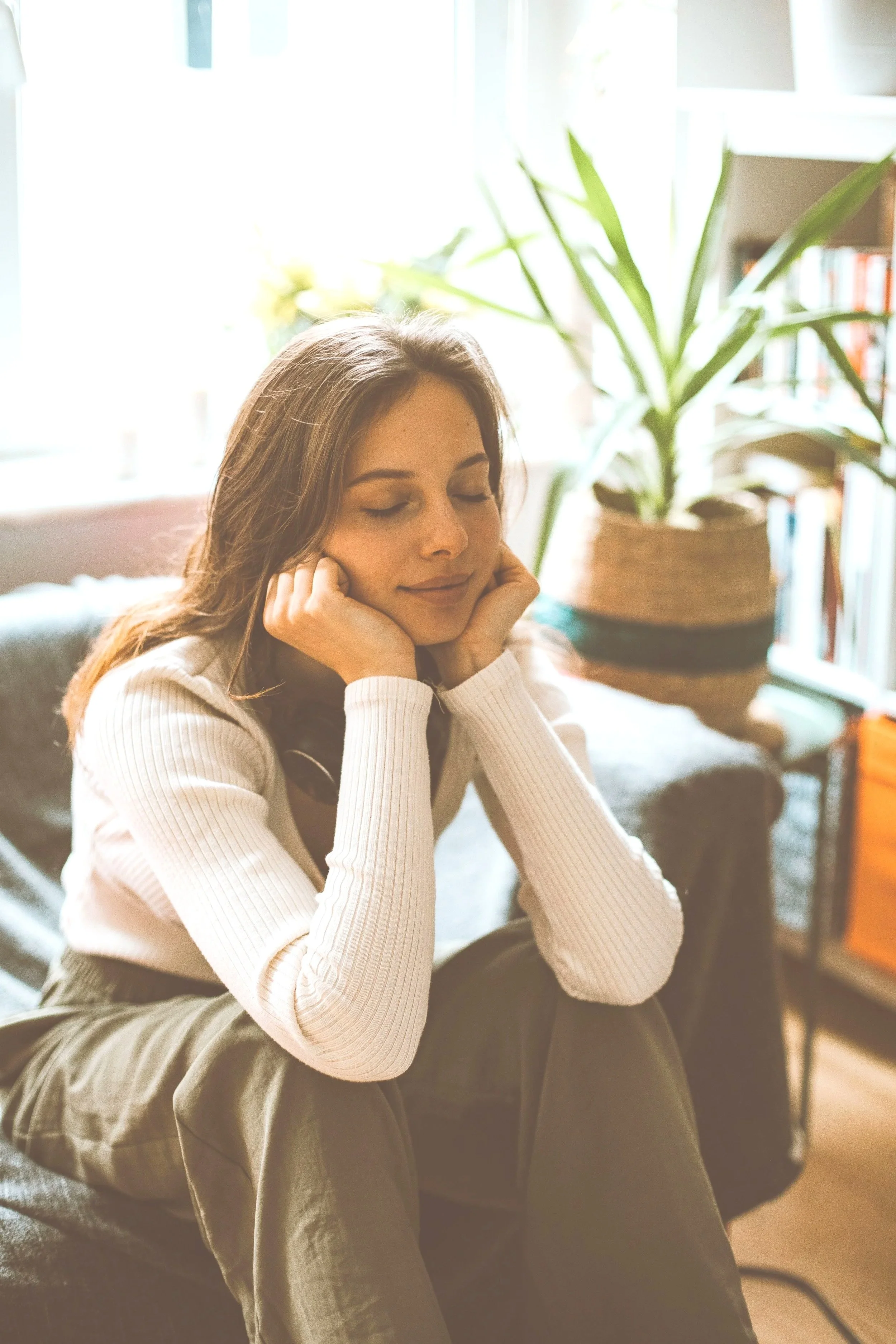 A young woman with long brown hair sits on a couch with her elbows on her knees and her hands on her cheeks, smiling with her eyes closed. She is wearing a white ribbed long sleeve shirt and olive green pants. Behind her, there's a large potted plant and a bookshelf in a well-lit room.