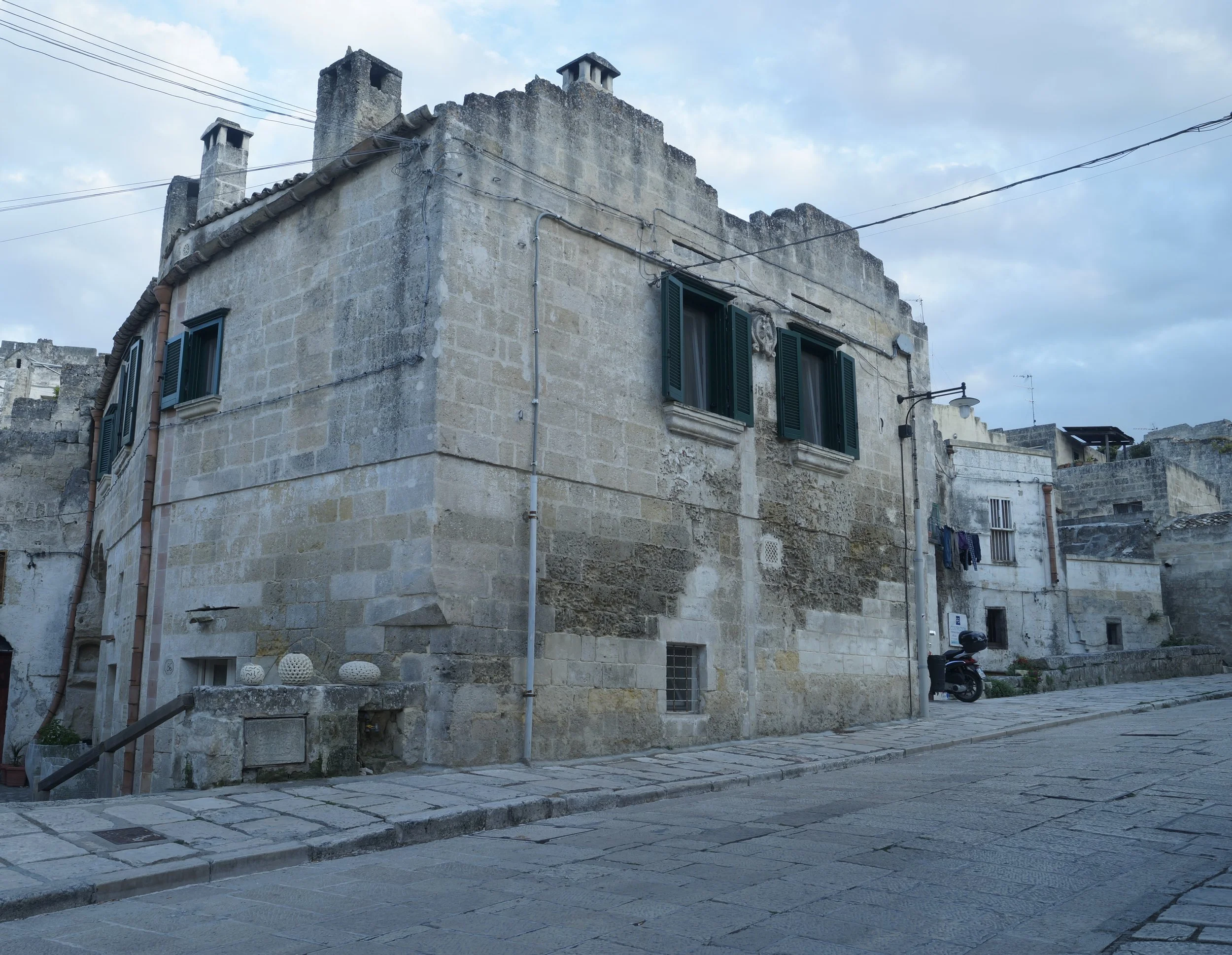 Vecchia casa in pietra con finestre verdi, in un quartiere tranquillo, strada di ciottoli e qualche scooter parcheggiato.