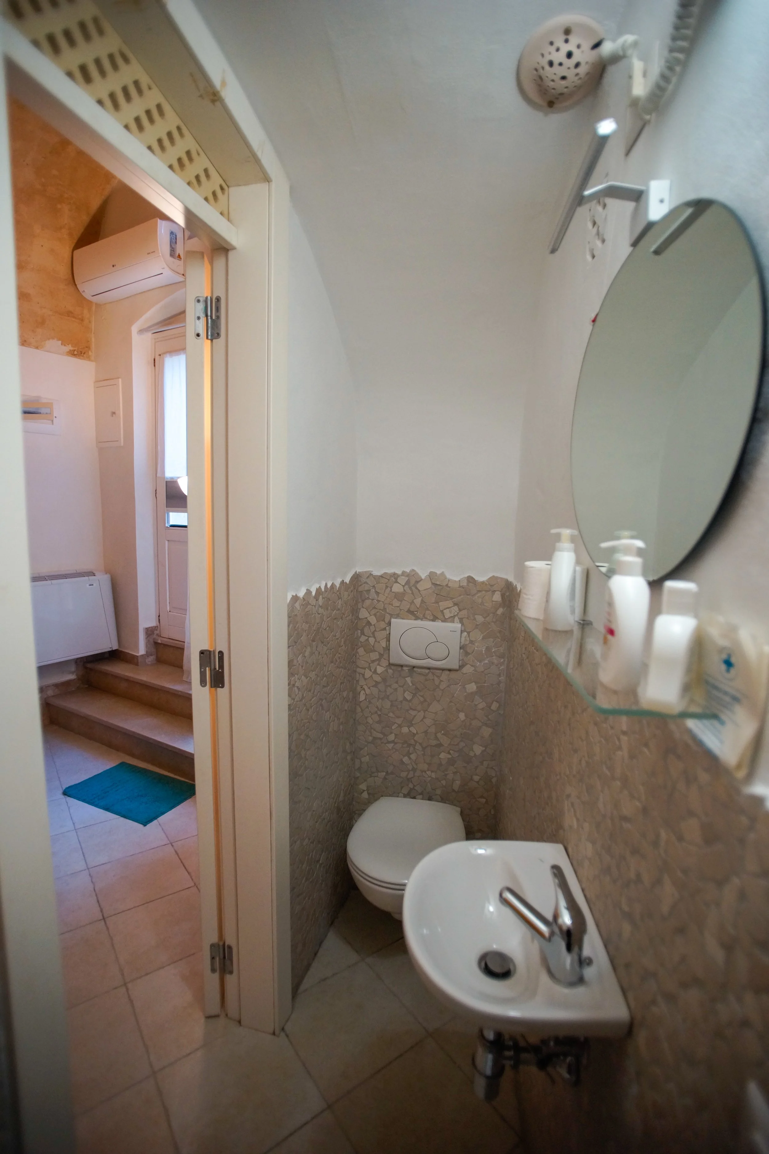 Piccolo bagno con lavabo, specchio rotondo, WC e pareti rivestite in pietra. Dispositivo di aerazione e alcuni prodotti per l'igiene personale sulla mensola.