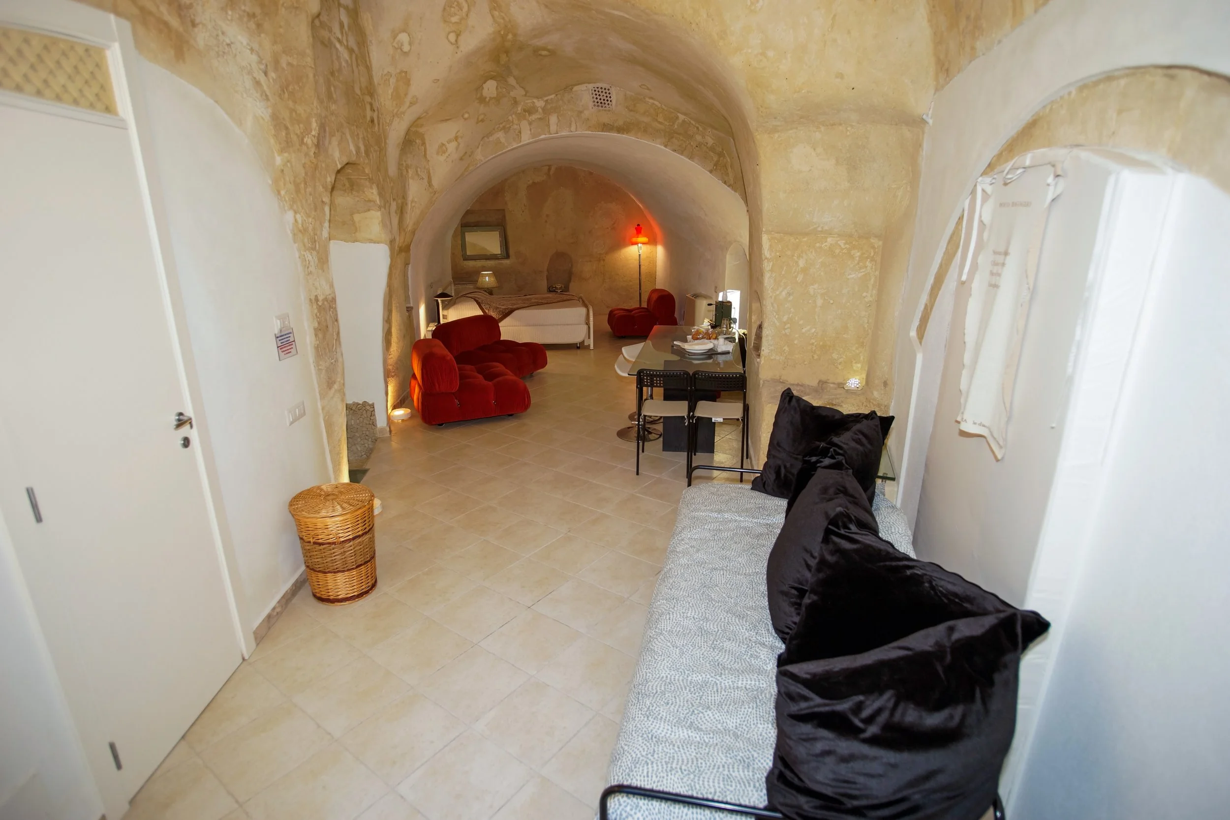 Interno di una stanza con pareti e soffitto in pietra, arredi moderni e da letto, divano nero, tavolo con cianfrusaglie, sedie nere, e poltrone rosse.