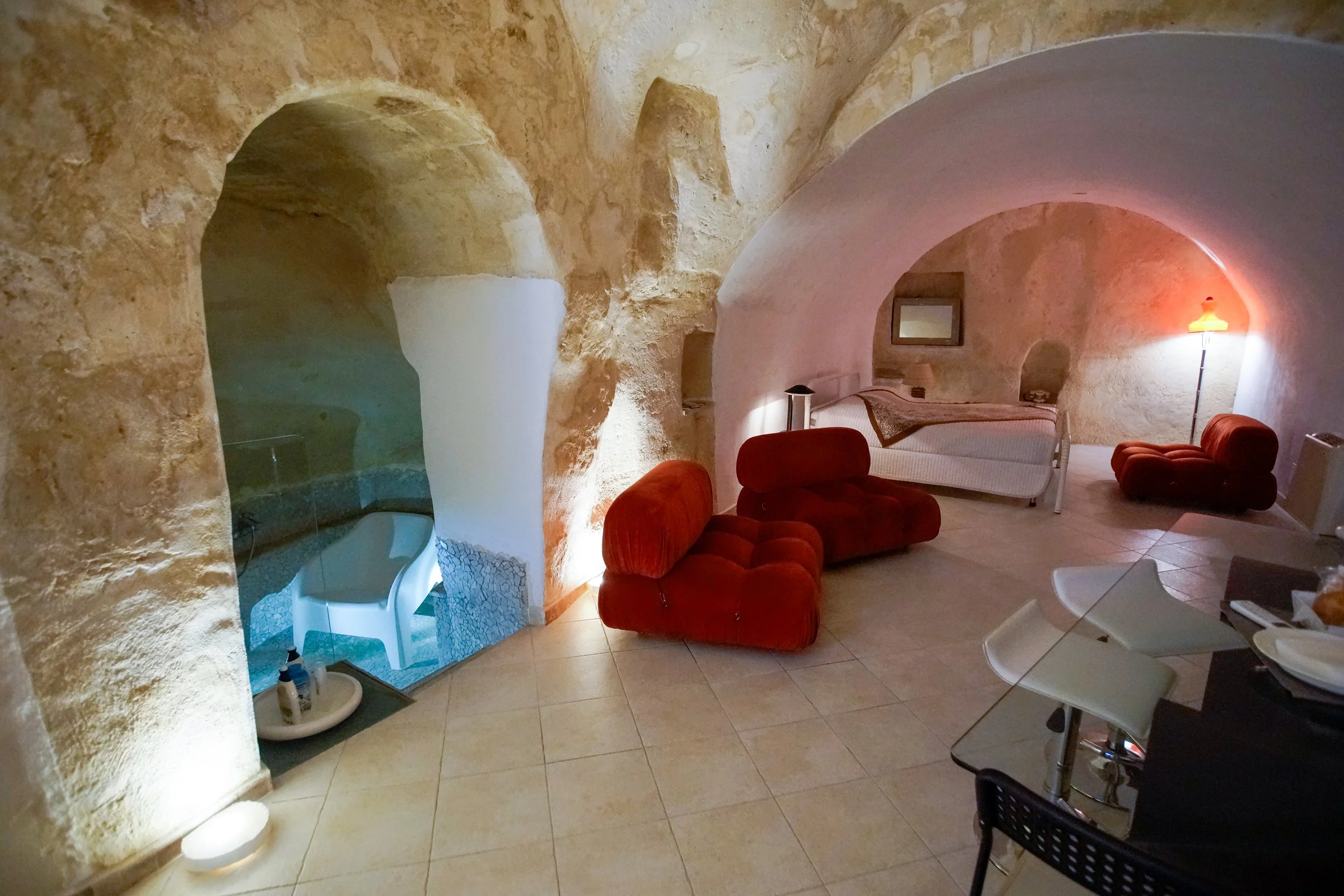 Interno di una stanza con pareti di roccia, letto, divani rossi e una sedia bianca