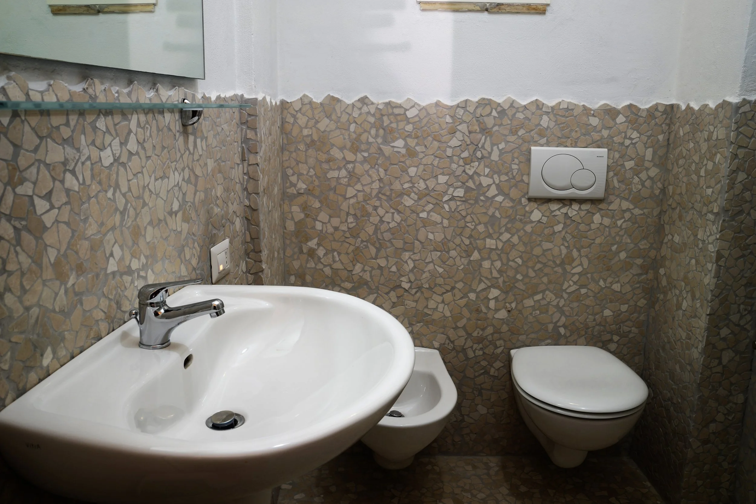 Bagno con sink bianco, bidet e WC sospeso, pareti rivestite con mosaico di pietra e una presa elettrica sotto uno specchio