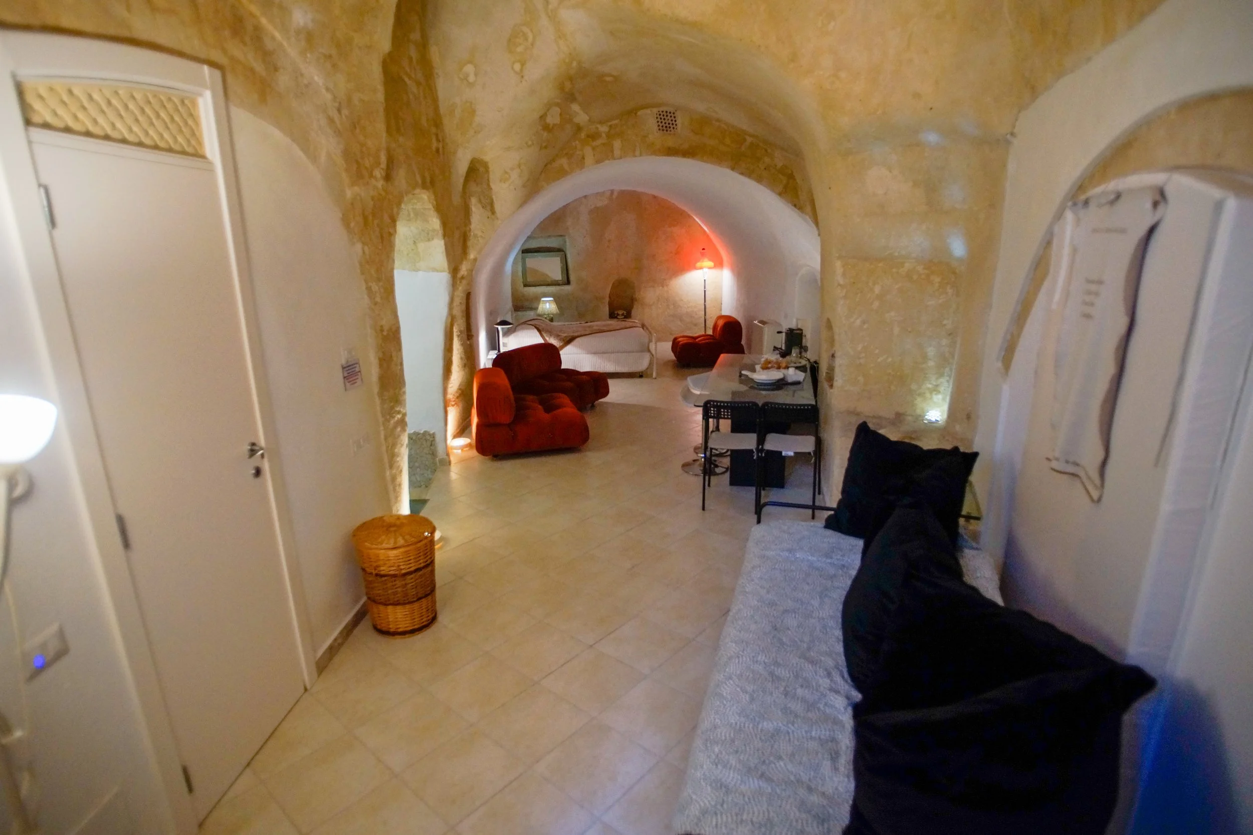 Interno di una stanza con pareti di tufo, divano nero, mobile con oggetti, due poltrone rosse, letto, lampade e un tavolo con cibo e bevande.