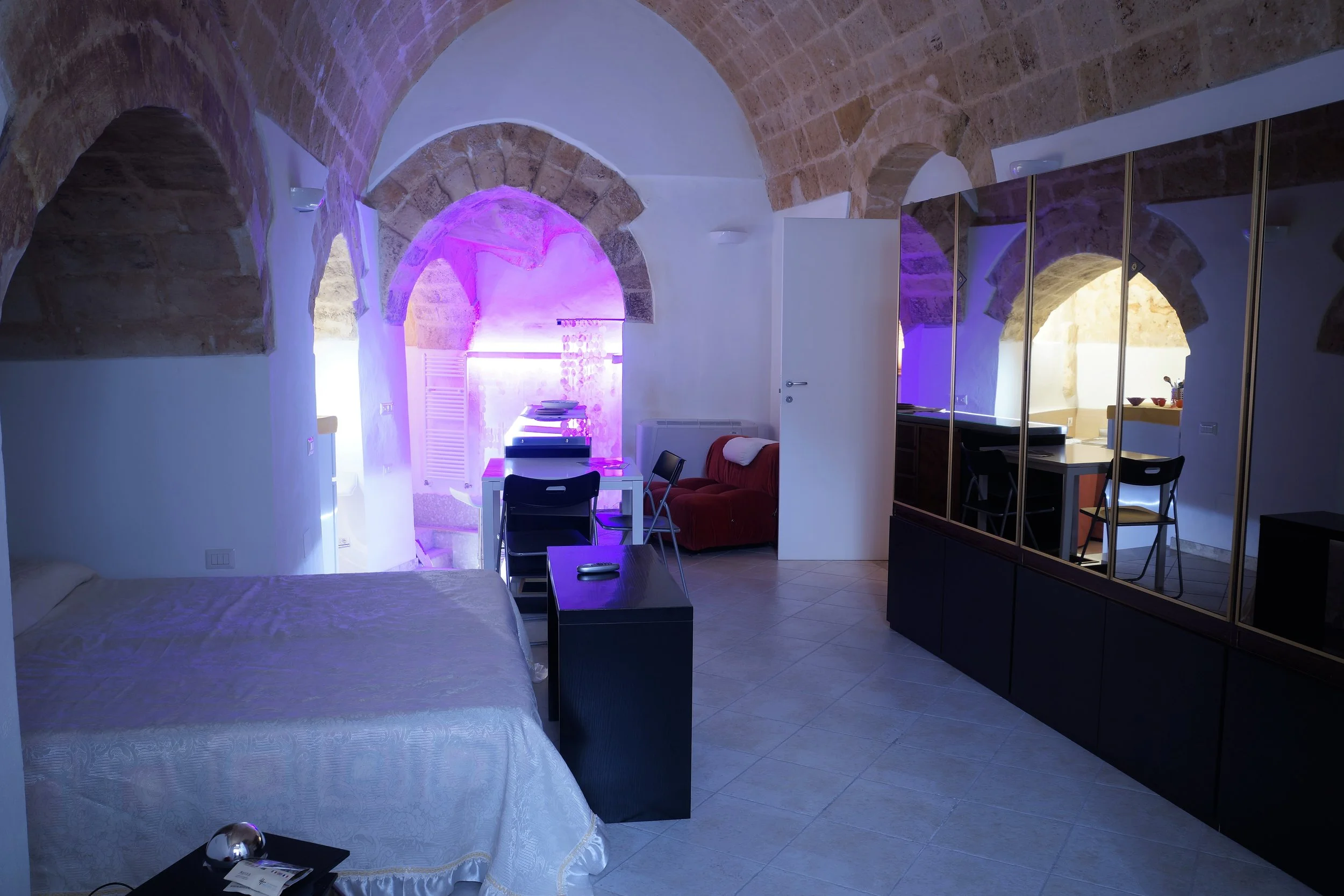 Interior di un appartamento con soffitti a volta in pietra, arredato con letto, divano rosso, tavolo, sedie e armadio con specchi, illuminato con luci viola e naturali