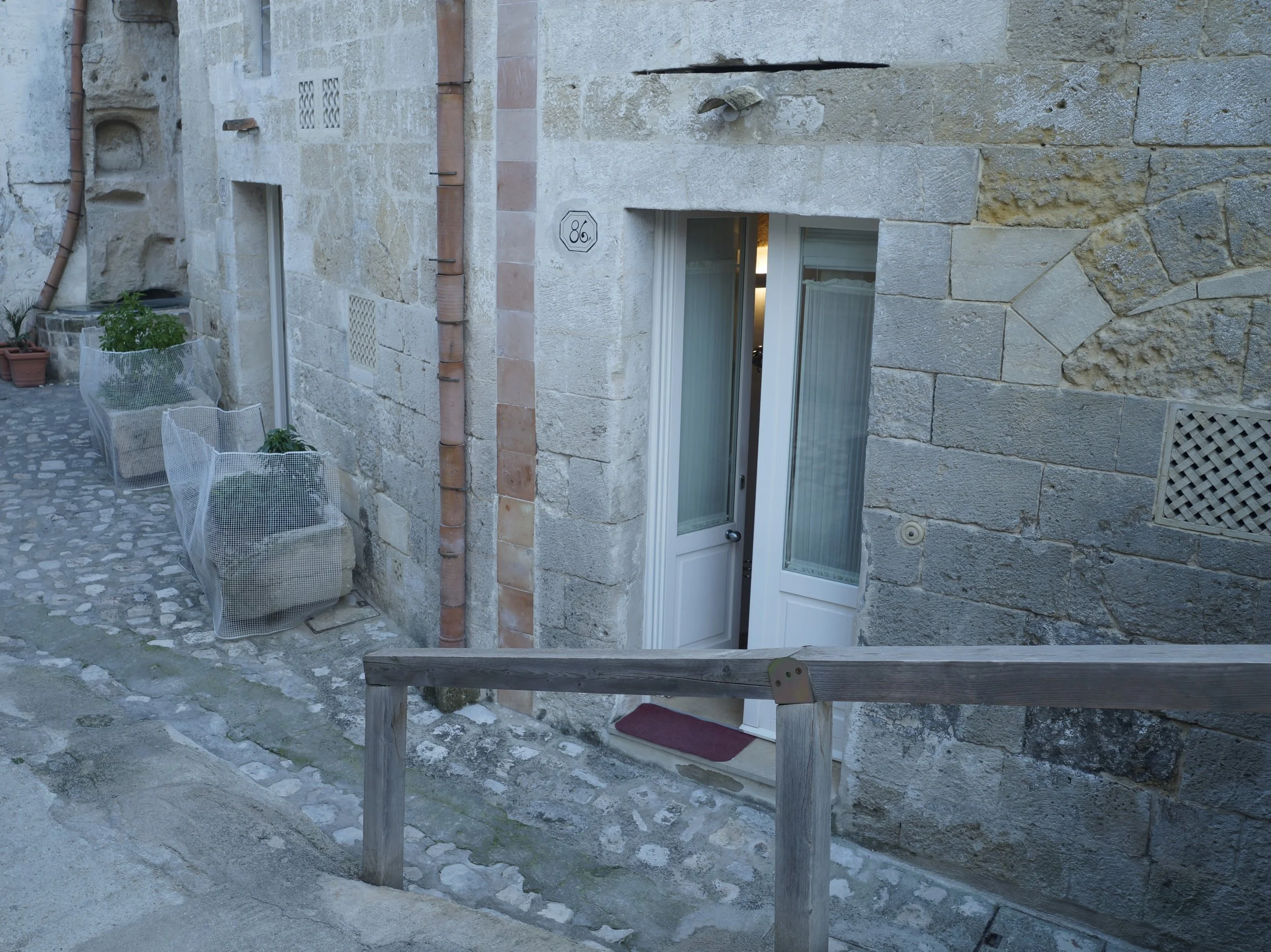 Facciata di un edificio in pietra con porta bianca, finestra e panchina con piante, pavimentata con pietre e cinta di legno.