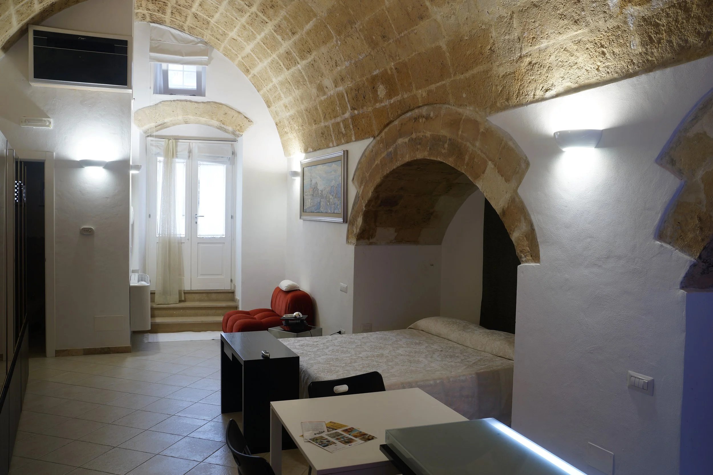 Interno di un appartamento con soffitti in pietra, divano rosso, letto, scrivania, tavolo con sedie, aria condizionata e accesso a un balcone con porta finestra.