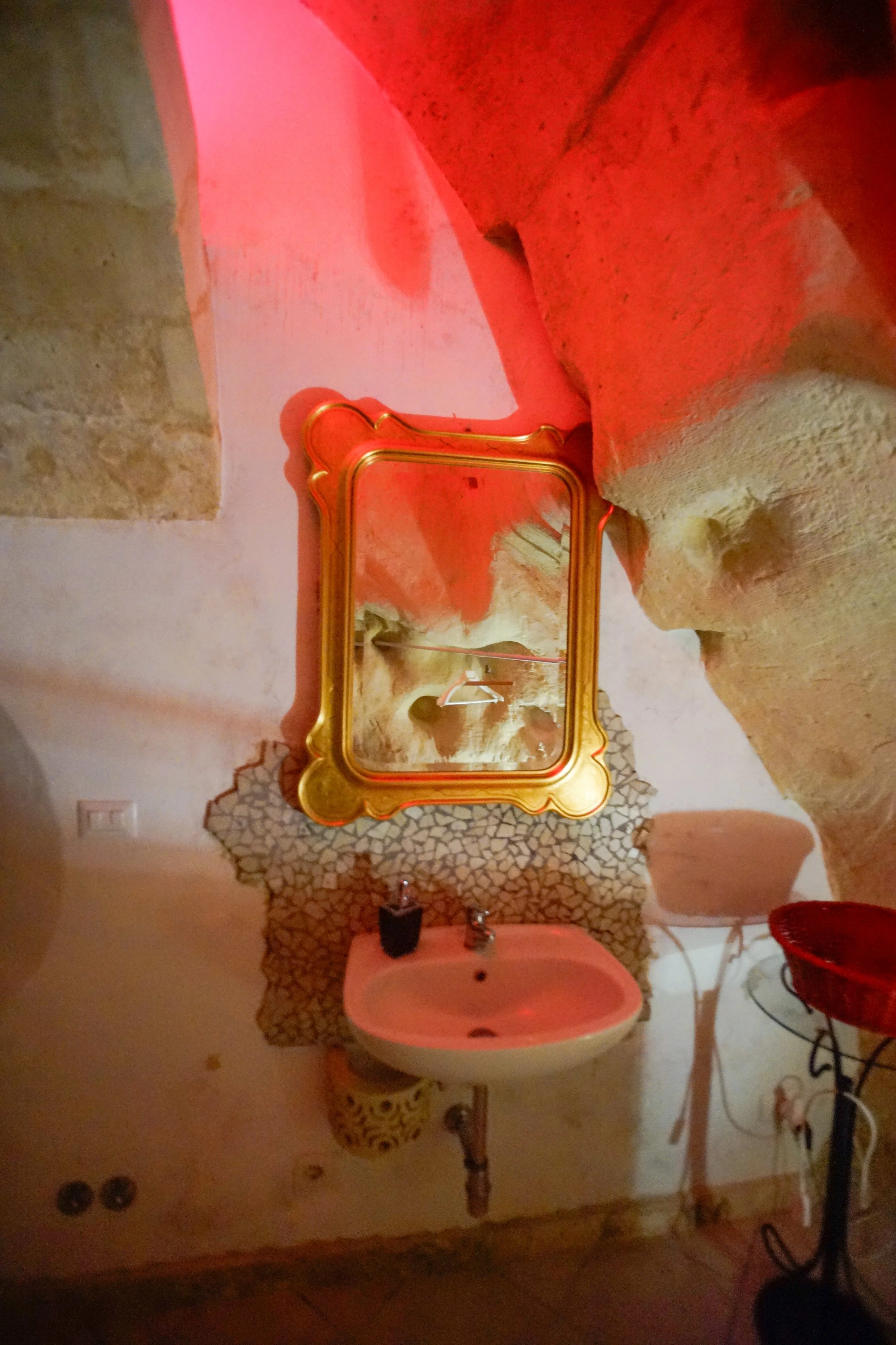 Specchio dorato sopra un lavabo con pareti in pietra e strutture in pietra, illuminato da luci rosse.