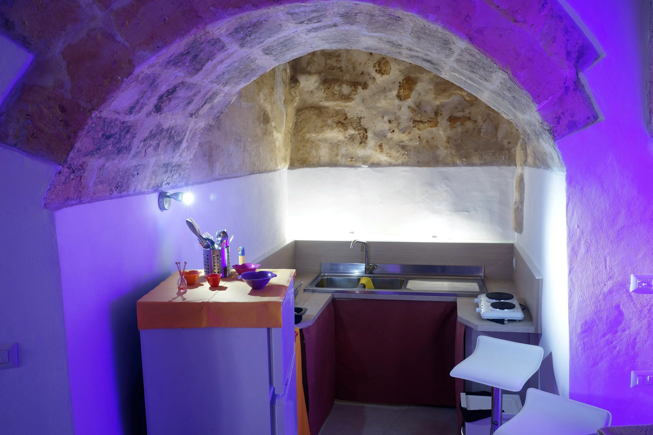 Piccola cucina con arco in pietra e luci blu e bianche, lavabo, due sedie bianche, piano cottura, e stoviglie colorate.