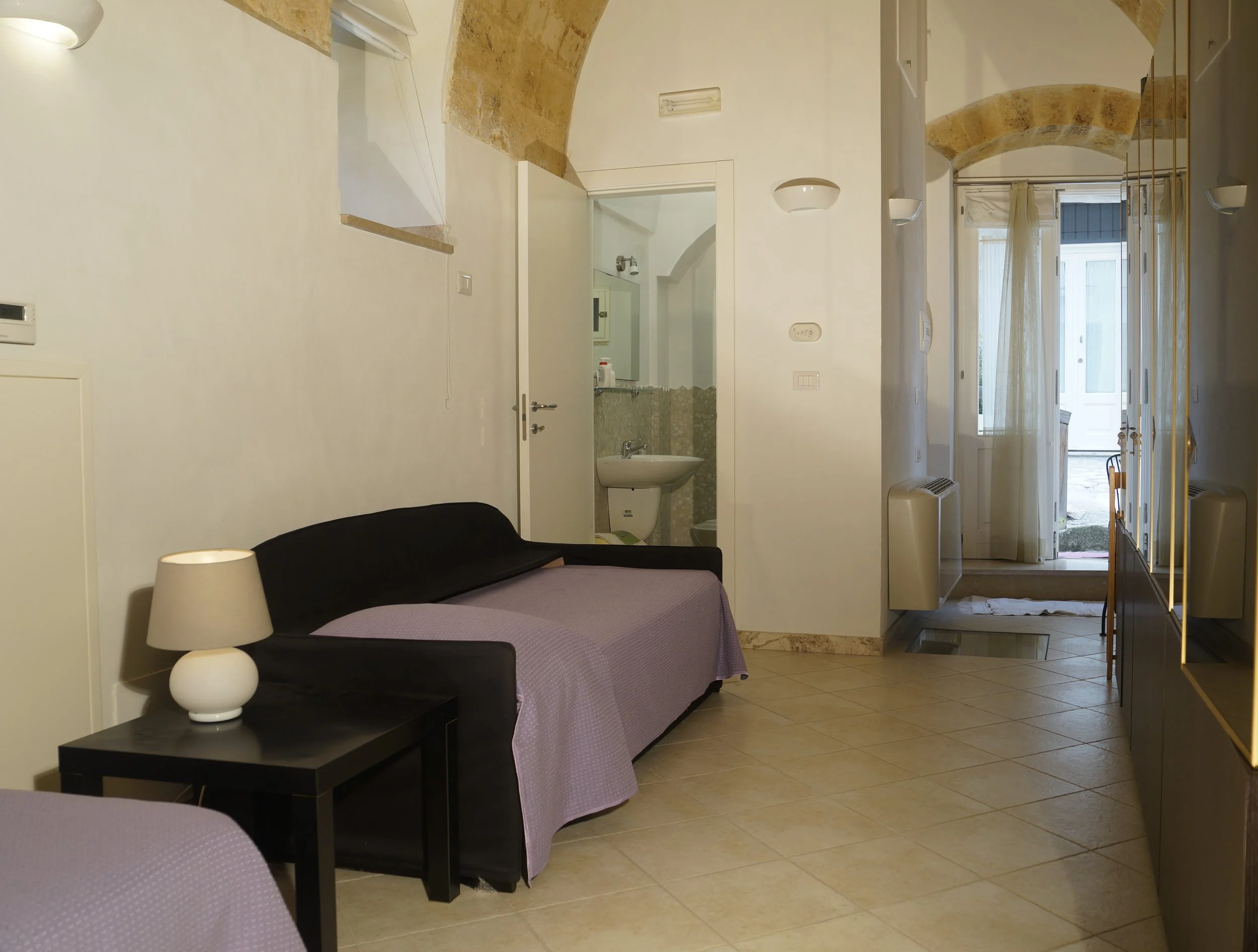 Interno di una stanza con divani, lampada, porta aperta su bagno e ingresso con porta finestra.