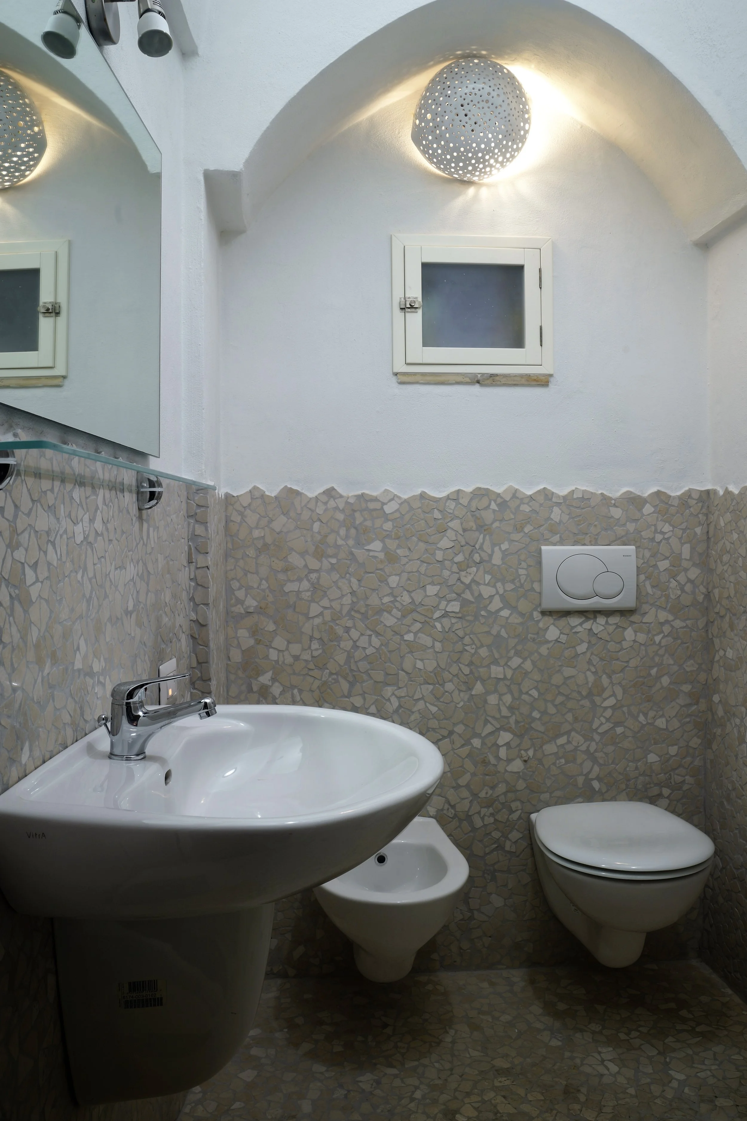 Bagno con lavabo, bidet e water, parete rivestita in mosaico beige, finestra con persiana, luce rotonda decorativa sulla parete.