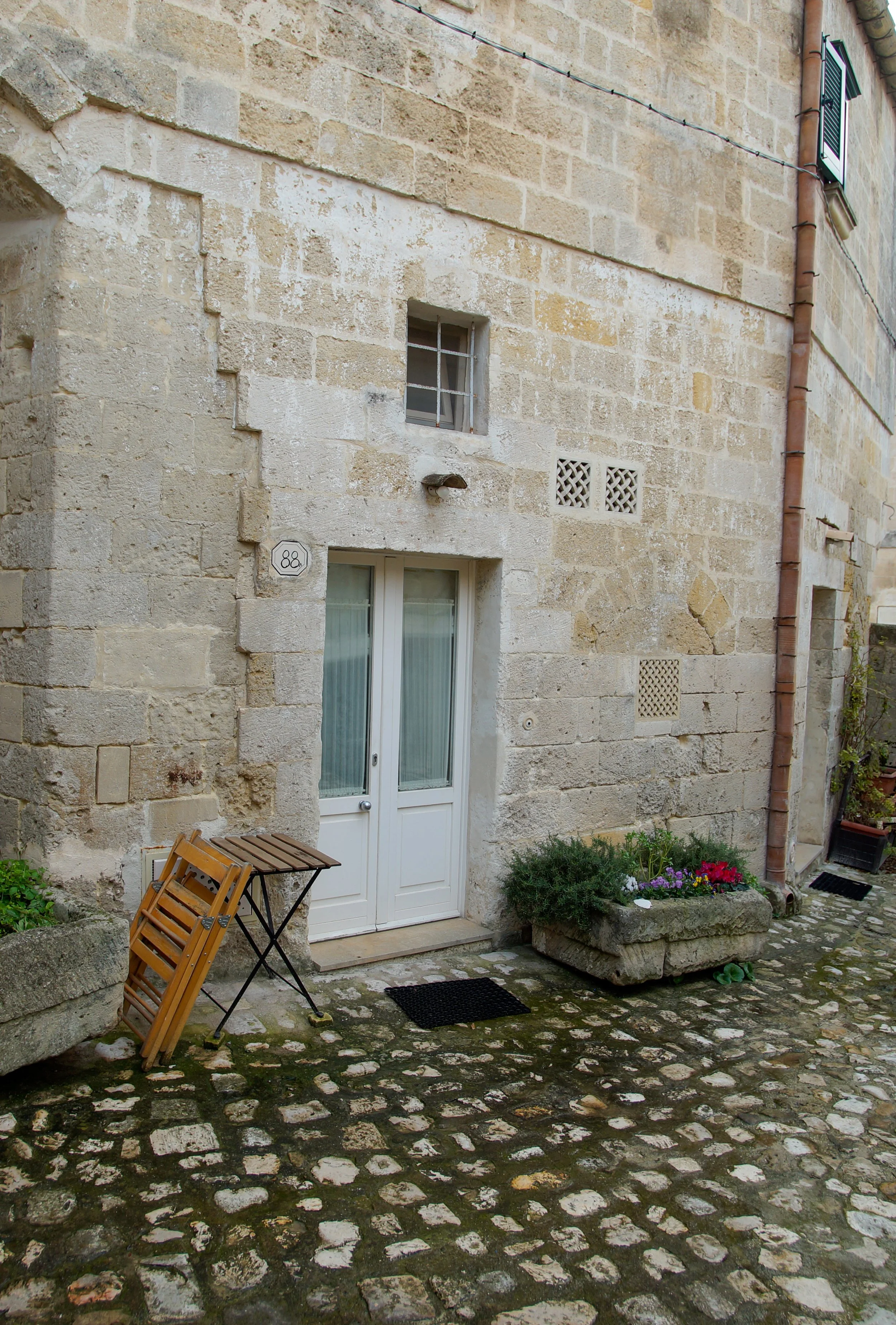Facciata di una casa in pietra con porta bianca, finestra con grate metalliche, piante fiorite in un vaso, tavolo di legno pieghevole, solette di pietra e un pavimento in ciottoli.