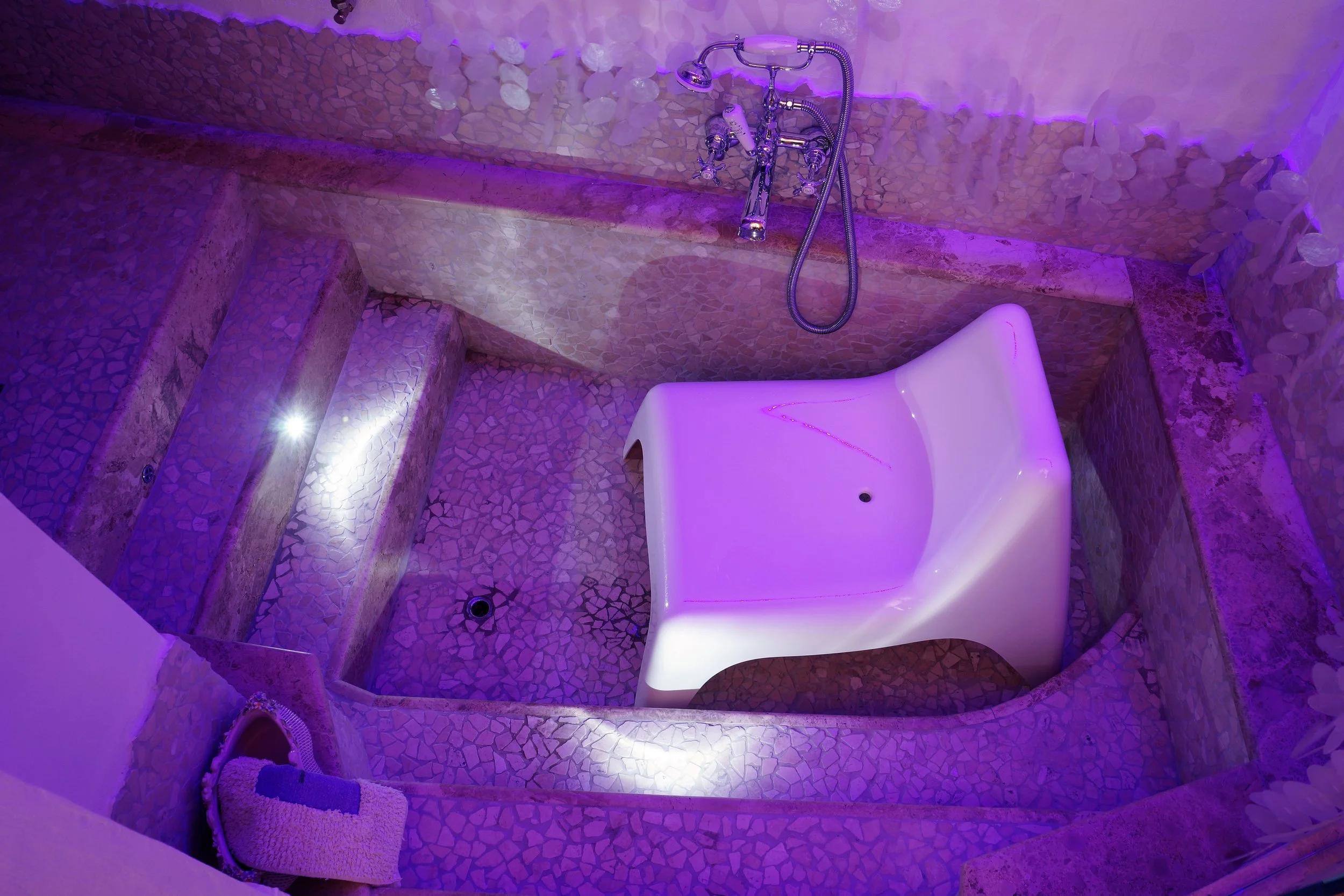 Vasca da bagno a forma di sedia, illuminata con luci rosa e viola, con pareti e pavimento rivestiti in mosaico di pietra, con fontana e doccia a parete