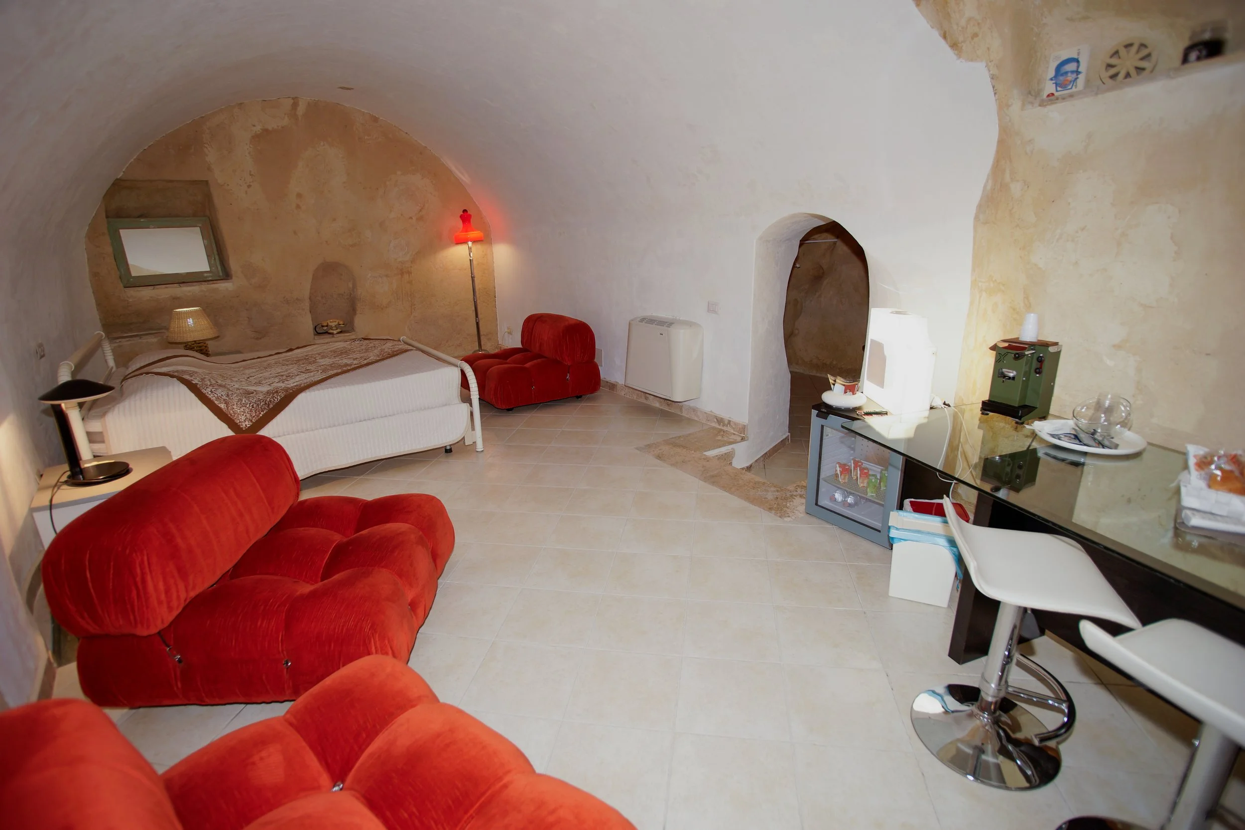 Interno di una stanza con soffitto a volta, letto, divano rosso, angolo bar, e piccoli elettrodomestici.